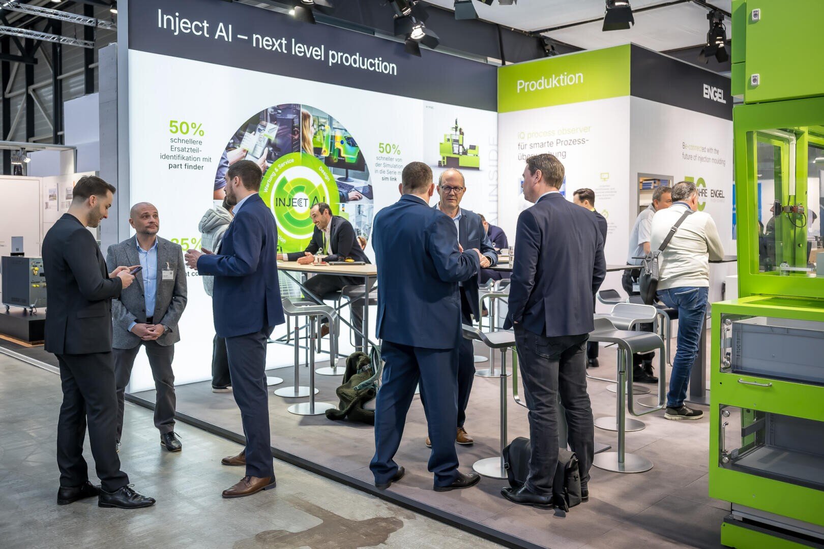 Un groupe de personnes en tenue d'affaires discute et établit des contacts à un stand d'exposition qui présente des affichages sur la production contrôlée par l'IA. Le stand est équipé de graphismes publicitaires, de sièges et d'équipements verts dans un hall d'exposition moderne.