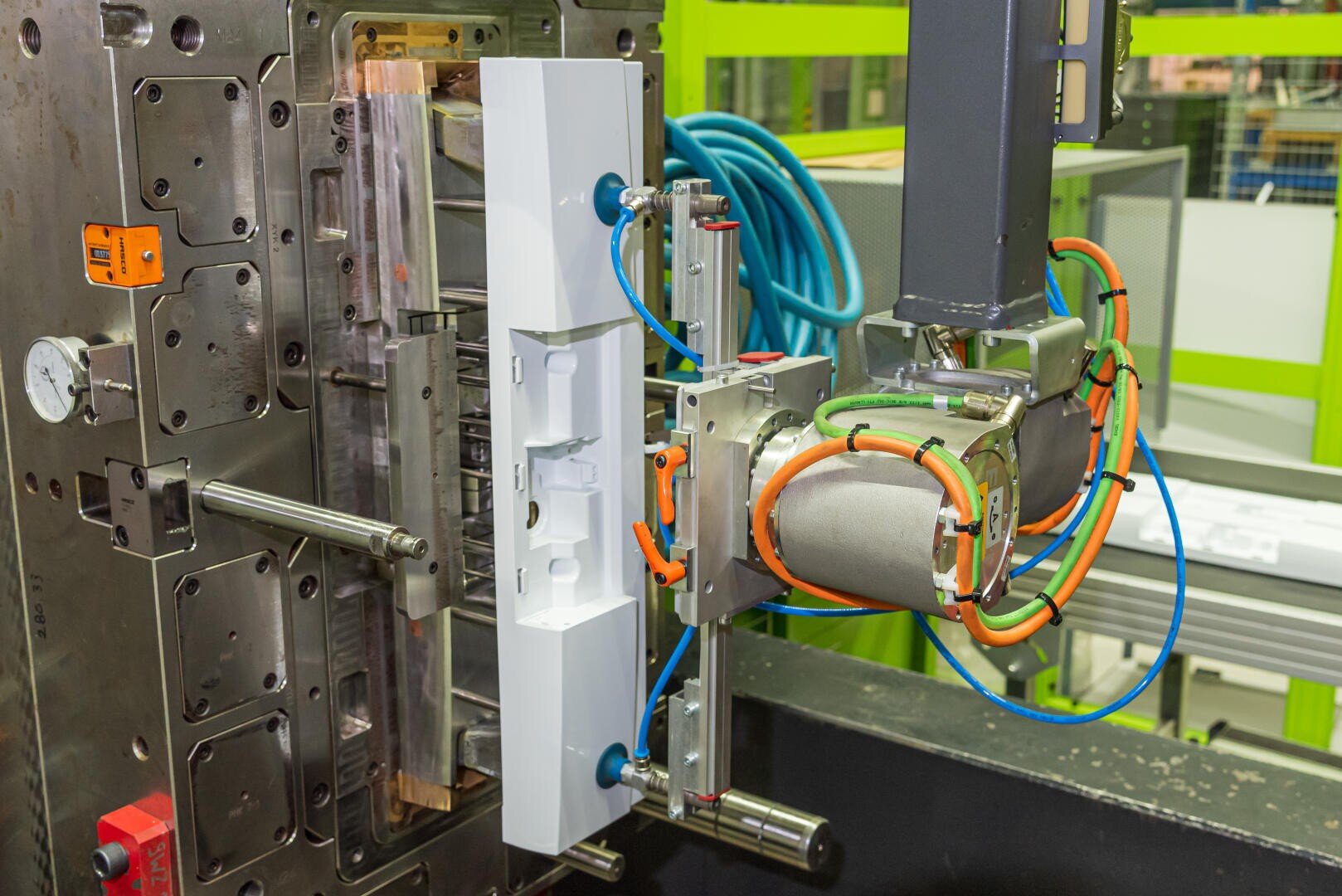 Ein Roboterarm mit orangefarbenen und blauen Kabeln entnimmt in einer industriellen Umgebung ein weißes Kunststoffteil aus einer Spritzgießmaschine.
