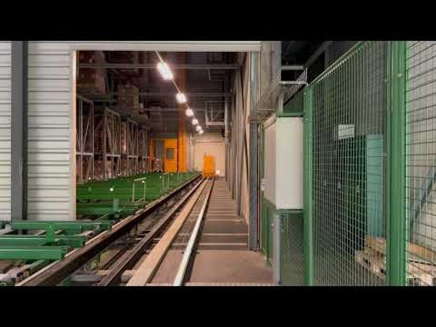 Langes, industrielles Lagerhaus mit Metallschienen, grünem Zaun und Oberlichtern. Am Ende der Gleise ist eine orangefarbene Tür zu sehen, an der linken Seite befinden sich Lagerregale und Geräte.