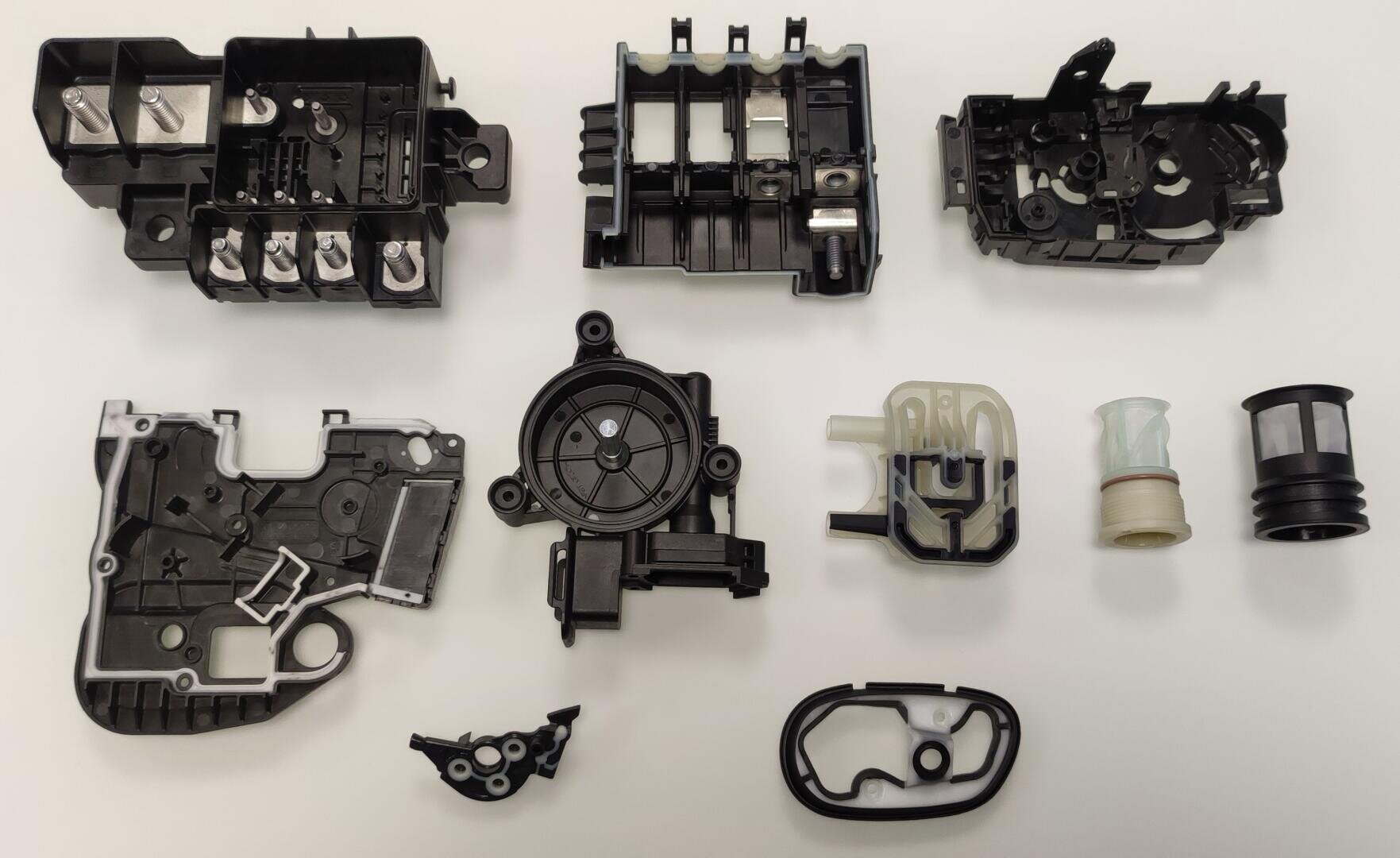 Verschiedene mechanische Teile aus Kunststoff und Metall, schwarz und grau, sind auf einer weißen Oberfläche angeordnet, darunter komplexe Gehäuse, Steckverbinder und zylindrische Bauteile.