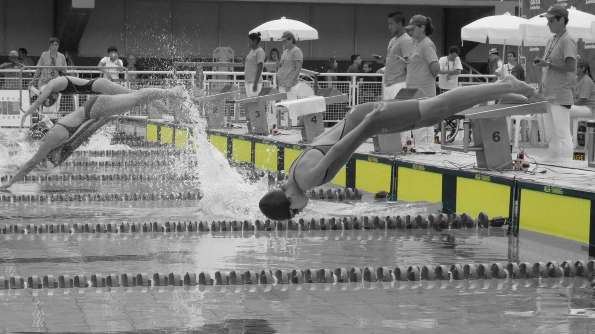 Schwimmer in Badeanzügen tauchen während des Starts eines Rückenschwimmwettbewerbs rückwärts in ein Becken. Es sind Wasserspritzer zu sehen, während Offizielle und Zuschauer vom Beckenrand aus zusehen. Das Bild ist überwiegend schwarz-weiß mit gelb hervorgehobenen Bahnmarkierungen.
