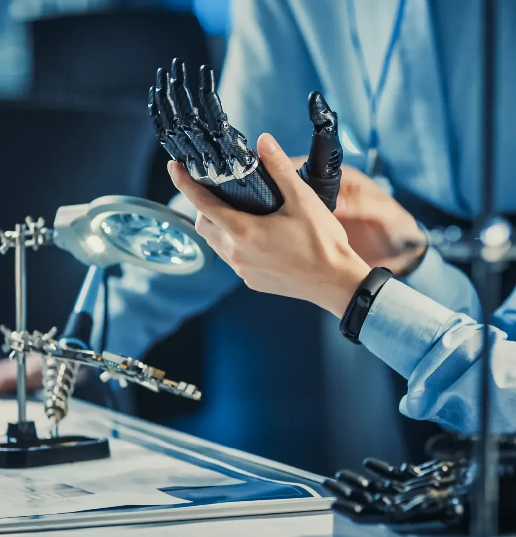 Eine Person in einem Laborkittel untersucht eine Roboterhand, die sie zur Inspektion auf einer Werkbank mit Werkzeugen, Plänen und mechanischen Bauteilen unter einer Schreibtischlampe hochhält.