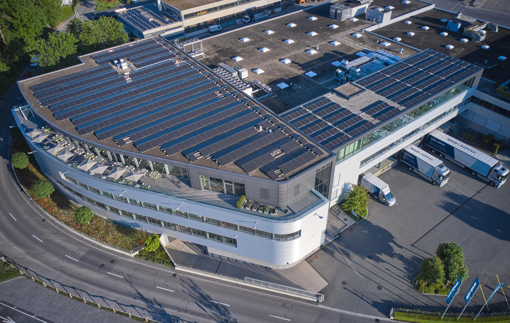 Luftaufnahme eines modernen Industriegebäudes mit Solarpaneelen, die einen Großteil des Daches bedecken, umgeben von Straßen, Grünflächen, geparkten Lastwagen und Laderampen. Das Gebäude hat eine geschwungene Fassade mit großen Fenstern.