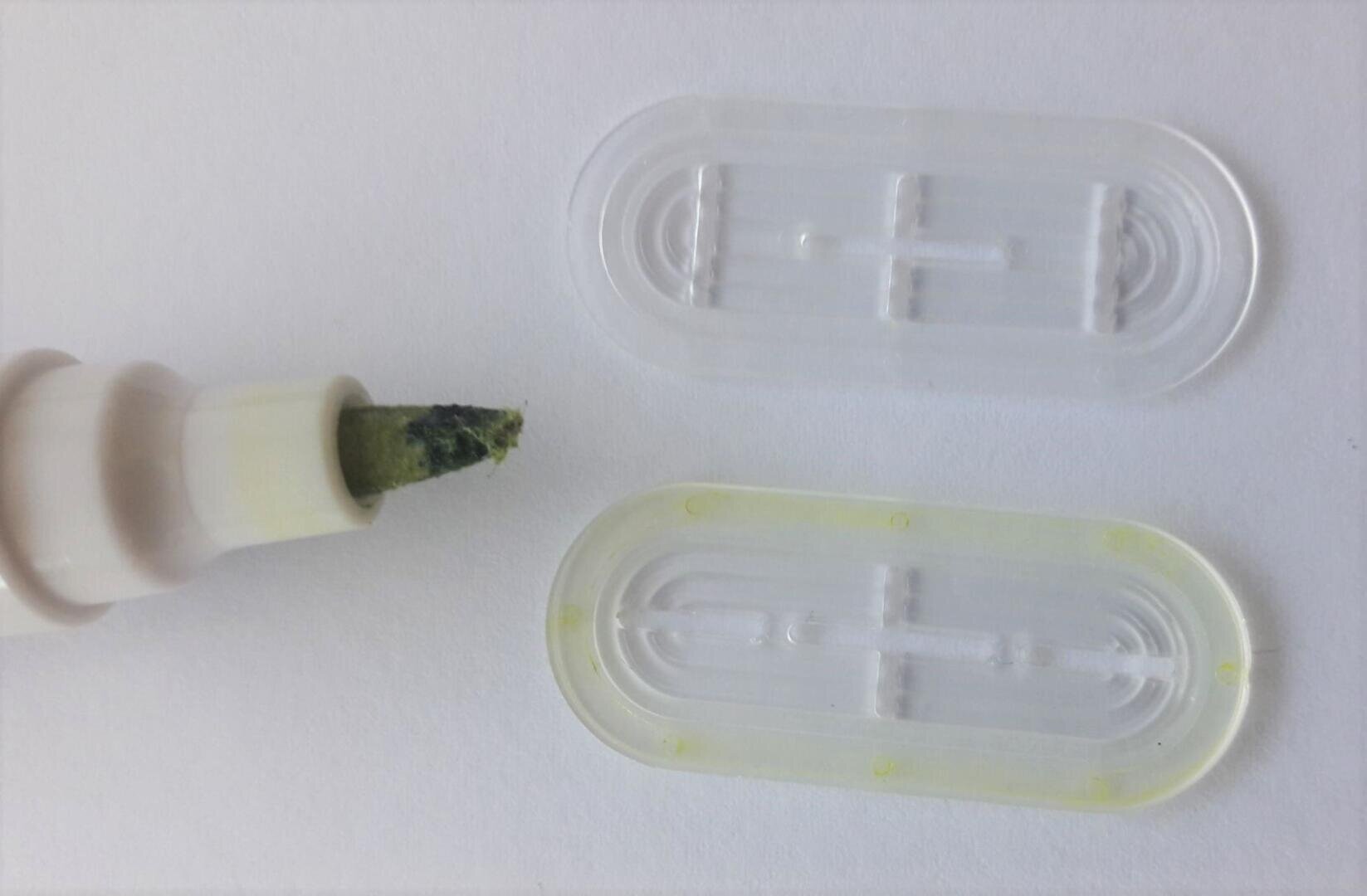 Nahaufnahme eines Markers mit freiliegender grüner Spitze neben zwei durchsichtigen Plastikkappen, die auf einer weißen Fläche liegen.