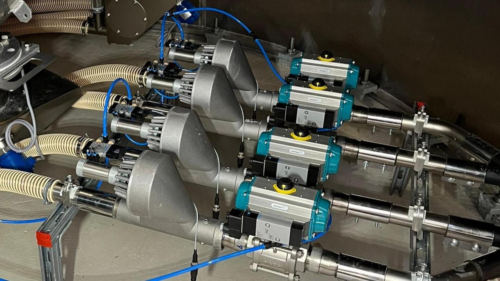 Vier industrielle pneumatische Stellantriebe mit blauen Komponenten sind auf Metallrohren montiert, die mit flexiblen Schläuchen und Kabeln verbunden sind und Teil eines mechanischen oder automatisierten Systems in Innenräumen sind.