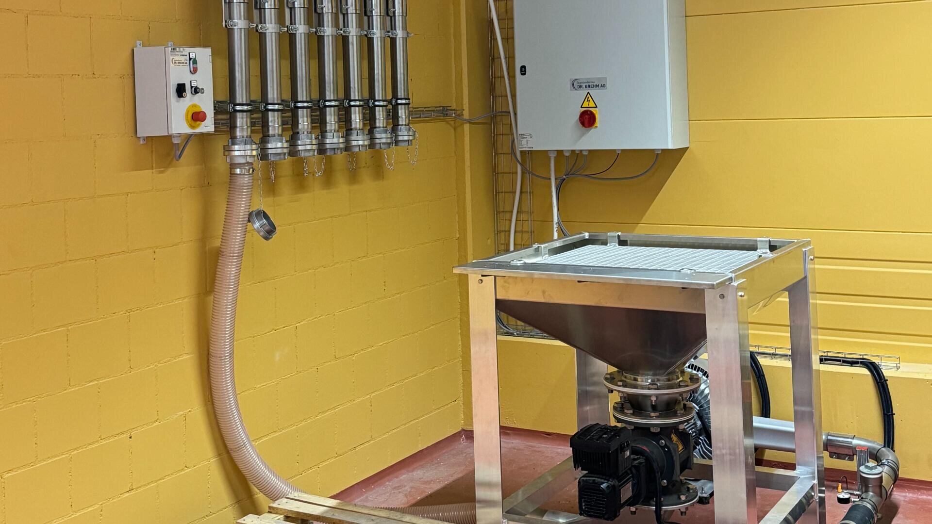 Industrielle Ausrüstung in einem Raum mit gelben Wänden, mit einem Metalltrichter mit Motor und einem Schlauch, der mit einer Tafel mit sechs vertikalen Rohren und Messgeräten verbunden ist, sowie einem an der Wand montierten elektrischen Schaltkasten.