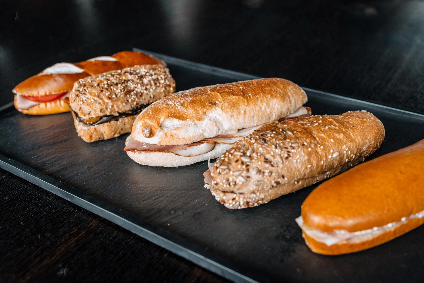 Eine Auswahl an verschiedenen Sandwiches mit unterschiedlichen Broten und Füllungen, die in einer Reihe auf einem dunklen rechteckigen Tablett mit dunklem Hintergrund angeordnet sind.