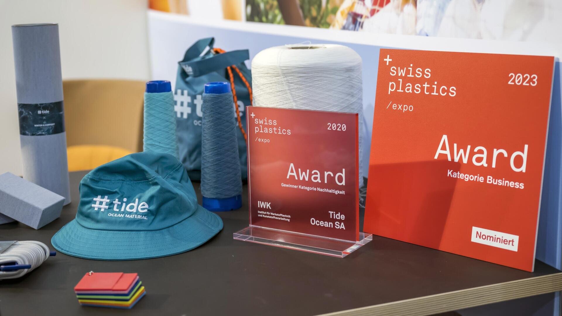 Une table d'exposition avec des badges Swiss Plastics Expo, une bobine de fil blanc, des bobines bleues, un chapeau #tide bleu-vert, des échantillons de tissus et des carrés colorés - le tout sur le thème de la durabilité des matières plastiques.