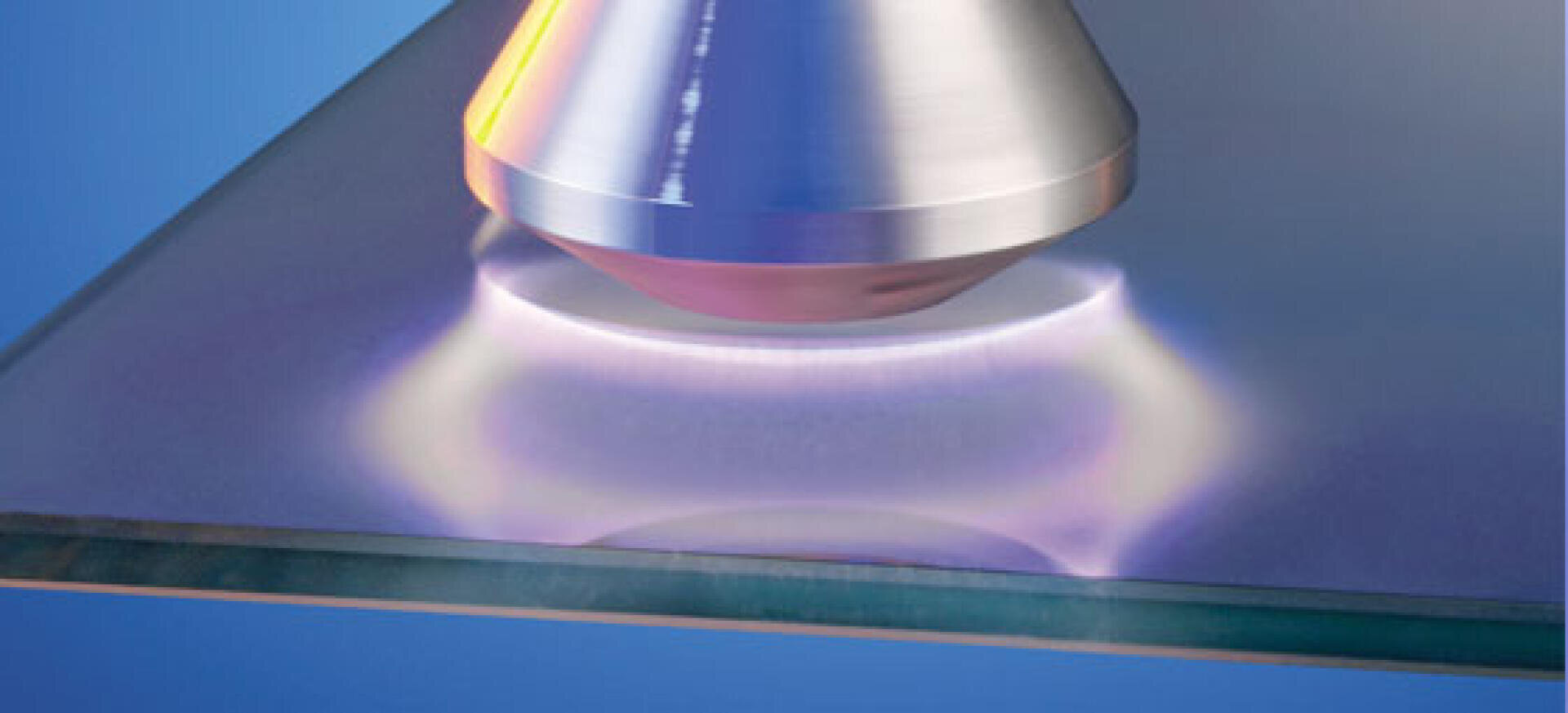 Gros plan sur une buse métallique émettant une douce lueur violette et bleue à la surface d'un matériau plat et transparent, probablement du verre, sur un fond bleu.