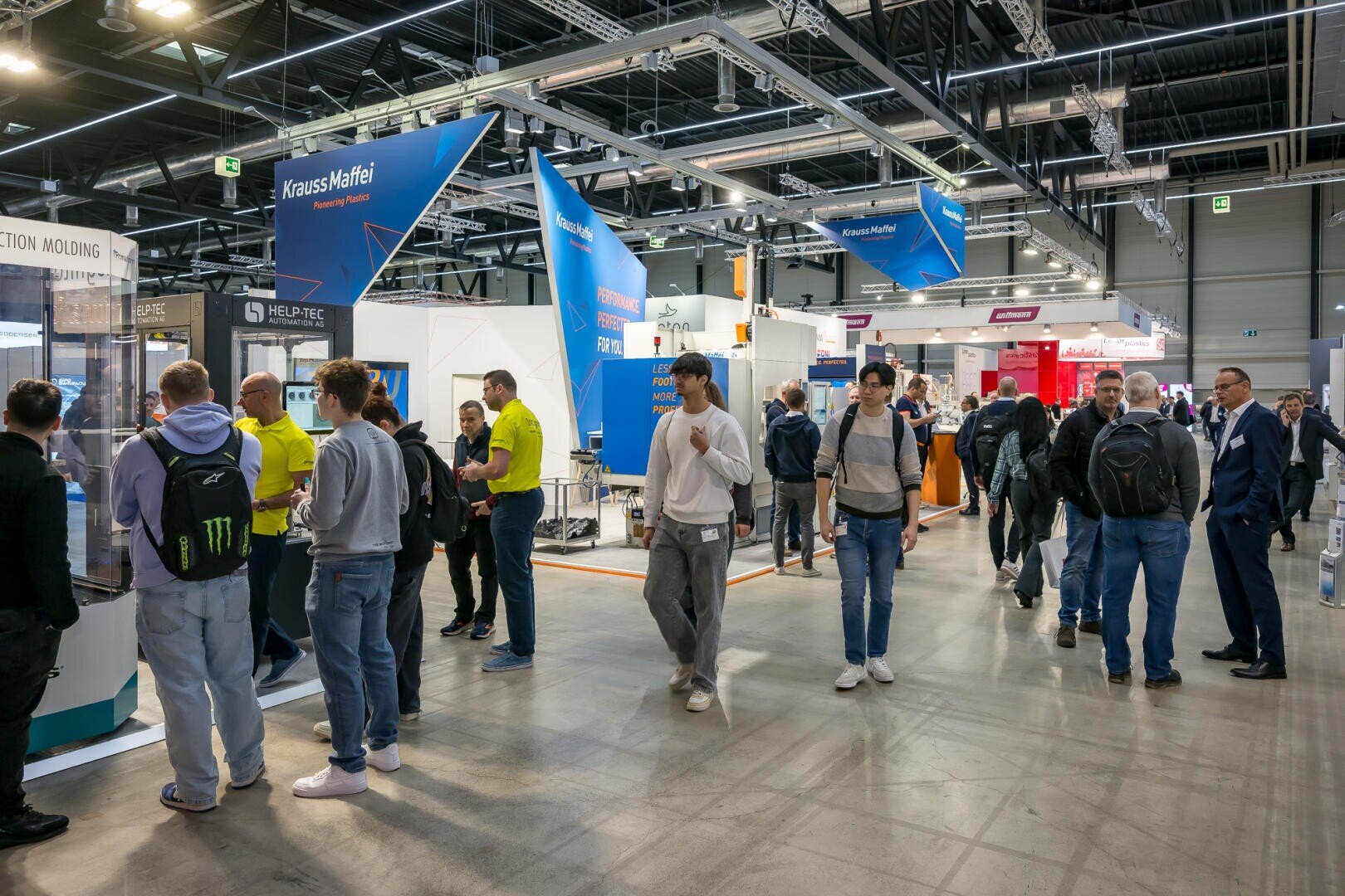 Auf einer belebten Indoor-Messe oder -Ausstellung laufen die Menschen um die verschiedenen Stände herum. Über den Ständen sind Schilder und Auslagen zu sehen, und es herrscht eine lebhafte Atmosphäre, in der die Teilnehmer die Veranstaltung erkunden.
