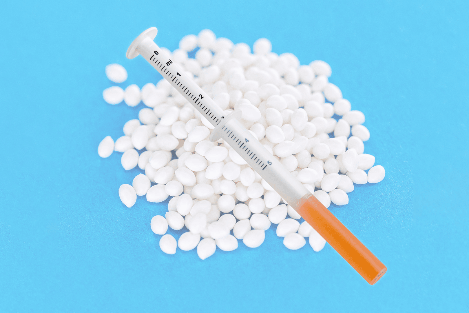 Eine mit orangefarbener Flüssigkeit gefüllte Spritze liegt auf einem Stapel kleiner, runder, weißer Pillen vor einem blauen Hintergrund.