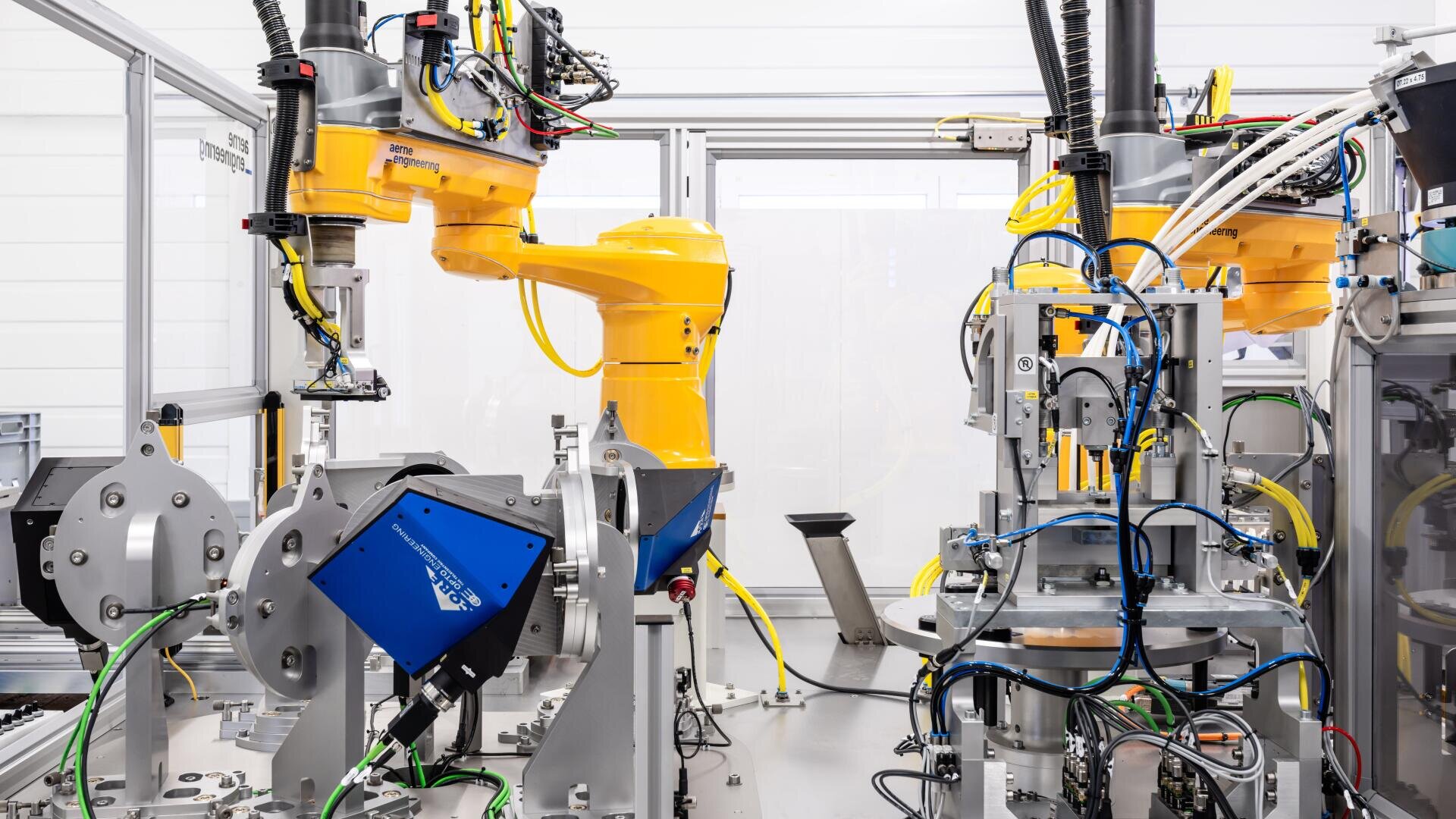 Deux bras robotisés jaunes travaillent dans un environnement industriel automatisé, entourés de différentes machines, câbles et appareils dans un environnement d'usine propre et moderne.