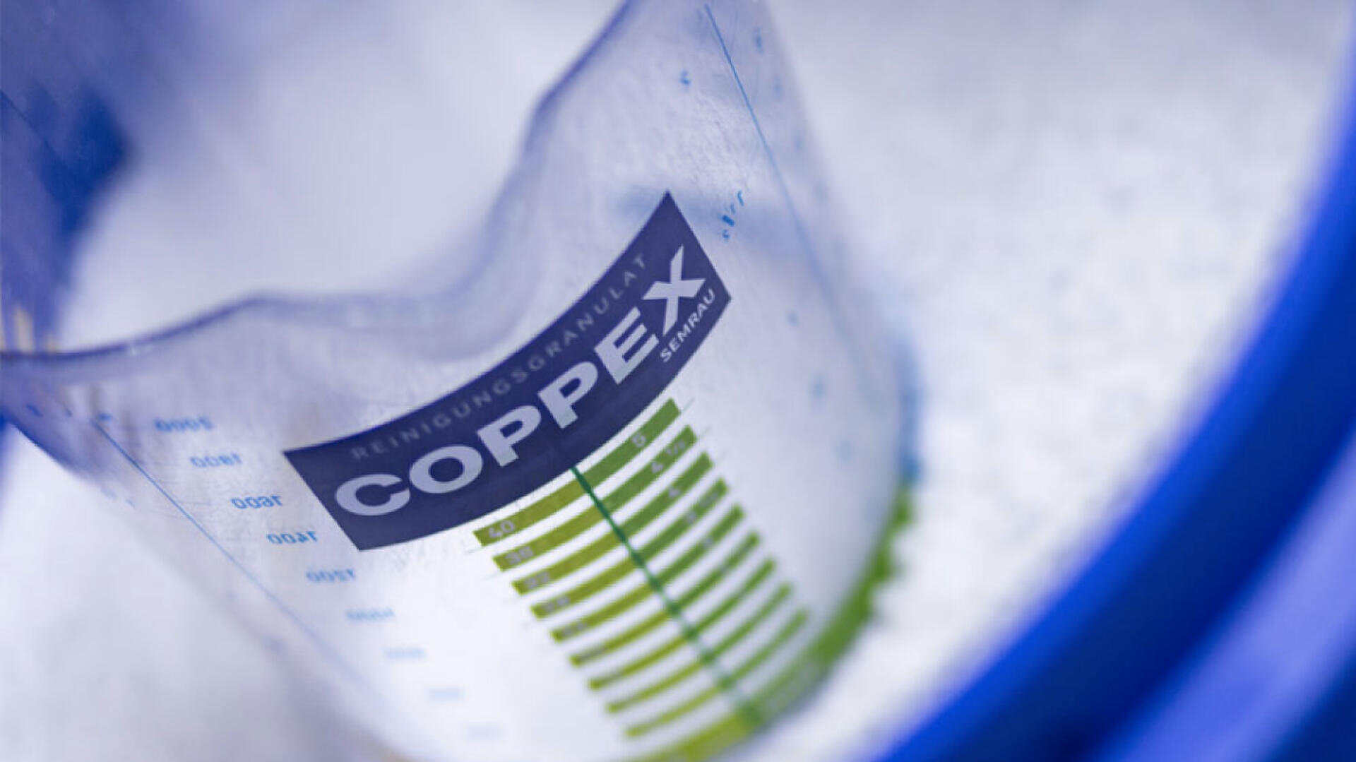 Nahaufnahme eines durchsichtigen Messbehälters mit der Aufschrift COPPEX und grünen und schwarzen Markierungen, der teilweise mit kleinen weißen Körnchen gefüllt ist. Der Behälter befindet sich in einem blauen, runden Rahmen.