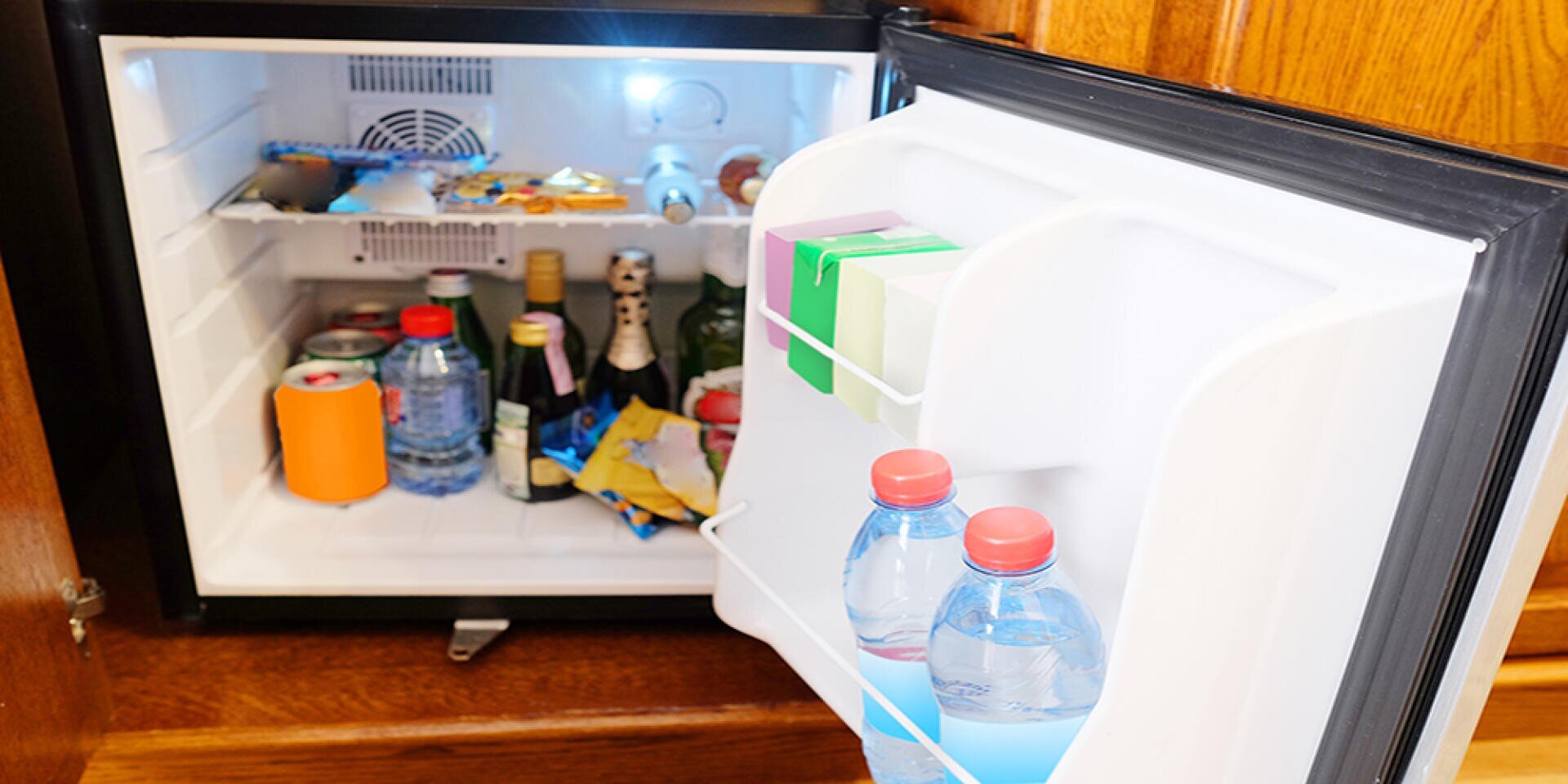 Ein offener Minikühlschrank mit verschiedenen Getränken, Snacks, Wasserflaschen und verpackten Artikeln auf den Regalen und in der Tür. Der Kühlschrank ist in einen Holzschrank eingebaut.