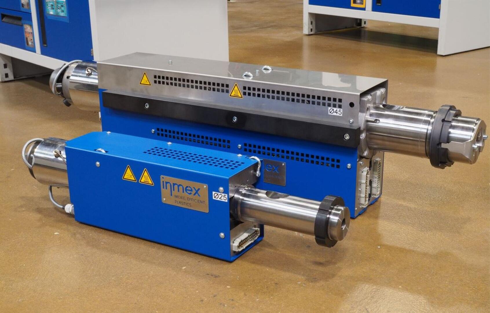 Deux machines industrielles bleues et argentées avec des pièces métalliques cylindriques et des symboles d'avertissement, placées sur un sol marron dans une usine ou un atelier. Les machines portent l'inscription inmex et sont équipées de grilles d'aération.