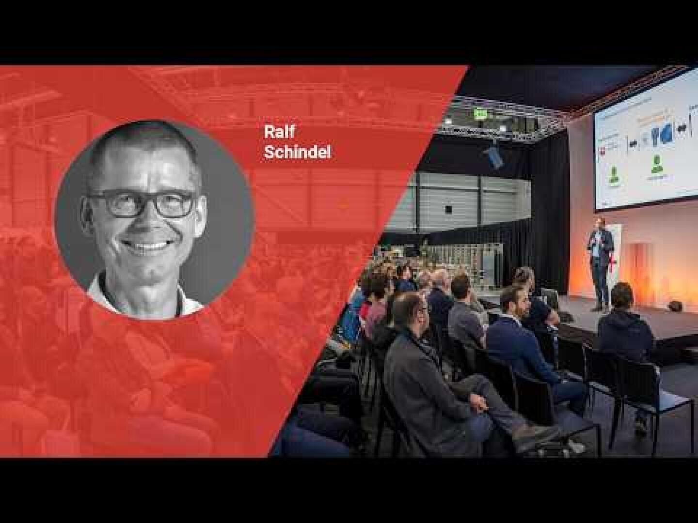 Eine Präsentationsveranstaltung mit einem sitzenden Publikum und einem Redner auf der Bühne. Auf der linken Seite zeigt ein rotes Grafik-Overlay ein Schwarz-Weiß-Kopfbild eines lächelnden Mannes mit der Aufschrift Ralf Schindel.