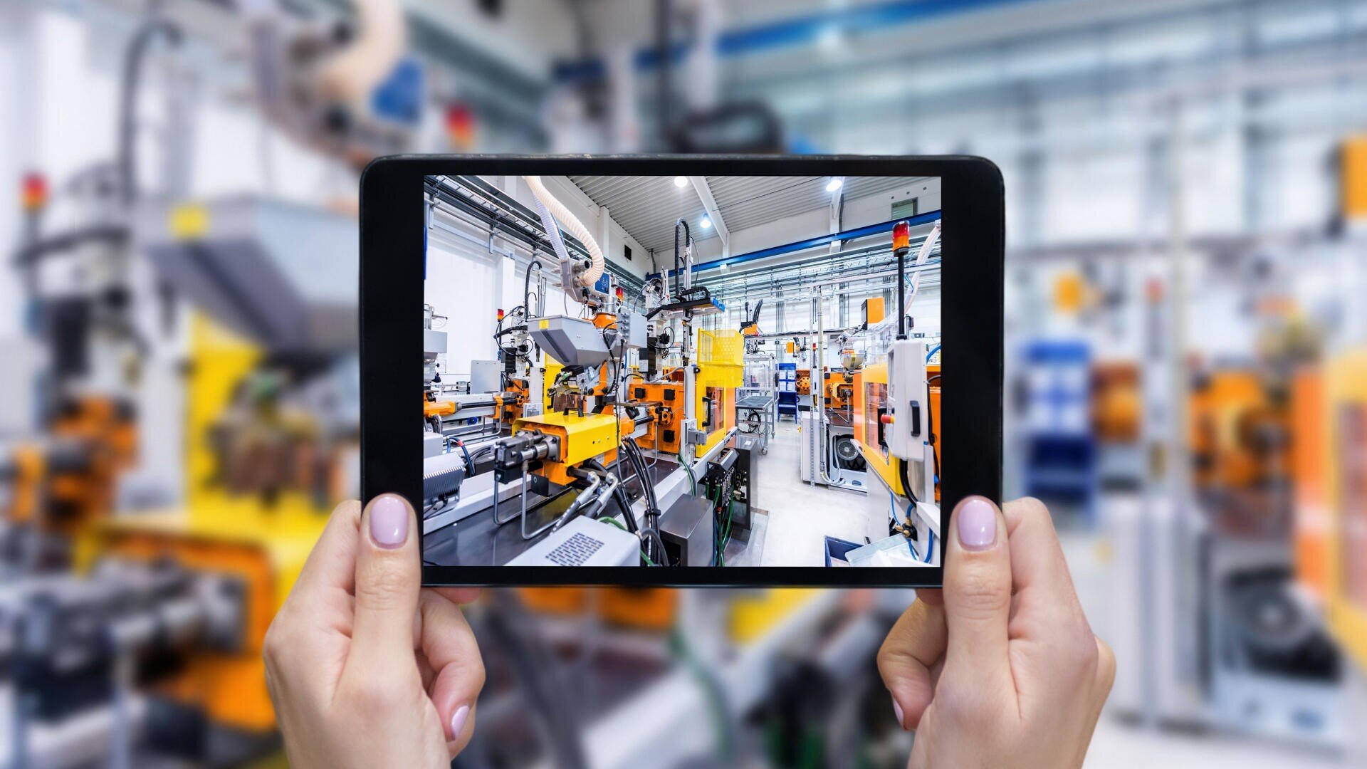 Eine Person hält ein Tablet in einer Fabrik und nimmt ein Bild von Industriemaschinen und -anlagen auf. Der Bildschirm des Tablets zeigt eine fokussierte Ansicht der Maschinen, während der Hintergrund unscharf erscheint.