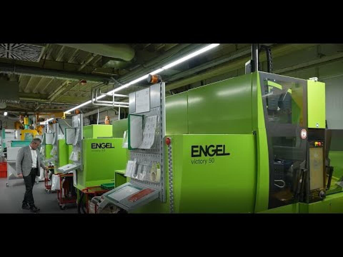 Ein Mann steht neben großen grünen ENGEL-Spritzgießmaschinen in einer hell erleuchteten Industrieanlage, an denen Papiere und Steuerungen angebracht sind.