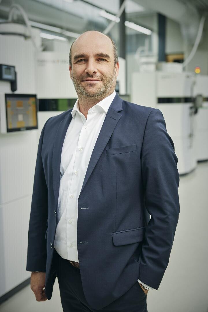 Un homme vêtu d'un costume bleu marine et d'une chemise blanche se tient debout, sûr de lui, dans un laboratoire moderne de haute technologie, avec des machines blanches et des écrans numériques en arrière-plan.