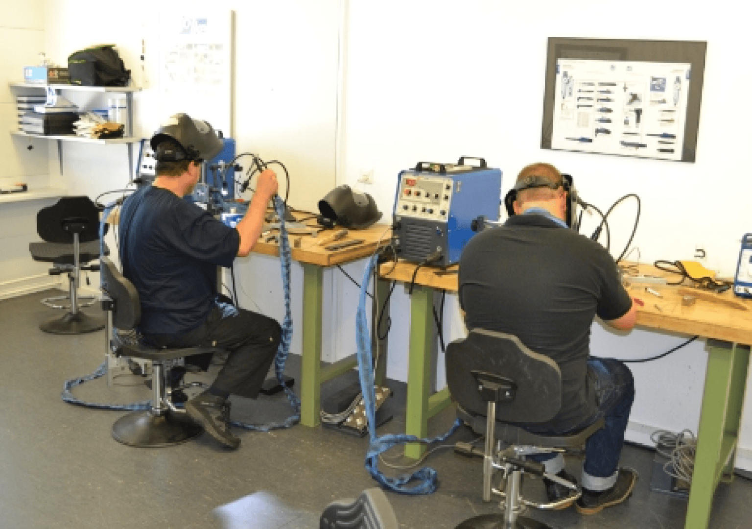 Zwei Personen sitzen an Werkbänken, tragen VR-Helme und halten Controller, mit denen sie das Schweißen in einer Werkstatt simulieren. Verschiedene Werkzeuge und Geräte befinden sich auf den Werkbänken und ein Diagramm ist an der Wand befestigt.