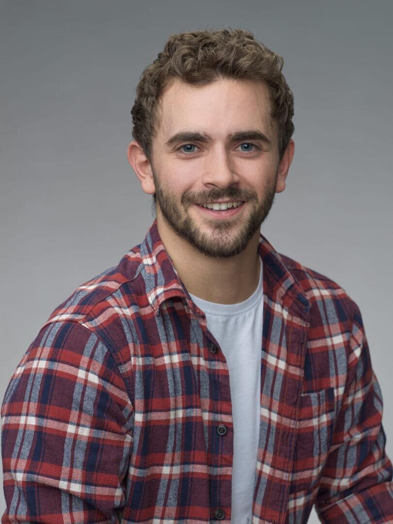Ein junger Mann mit lockigen braunen Haaren und einem Bart lächelt in die Kamera. Er trägt ein rot-blau kariertes Hemd über einem weißen T-Shirt vor einem schlichten grauen Hintergrund.