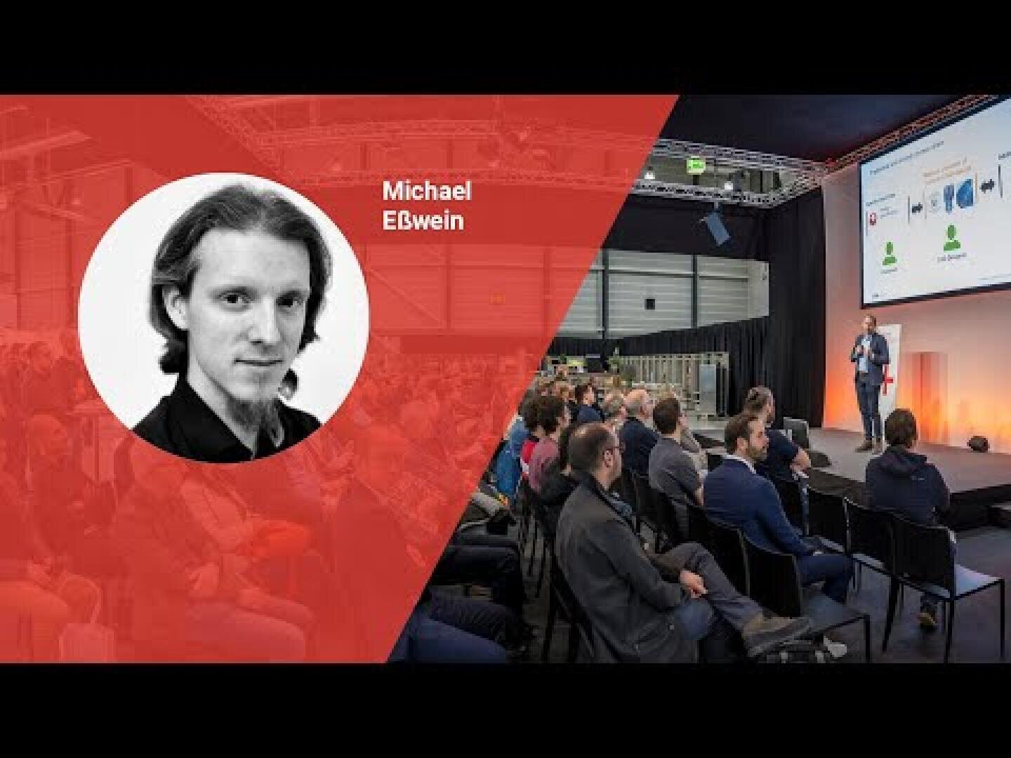 Ein Redner spricht in einem modernen Konferenzraum zu einem Publikum. Auf der linken Seite zeigt ein roter Bildschirm ein Schwarz-Weiß-Porträt von Michael Eiswein. Ein großer Bildschirm zeigt eine visuelle Präsentation.