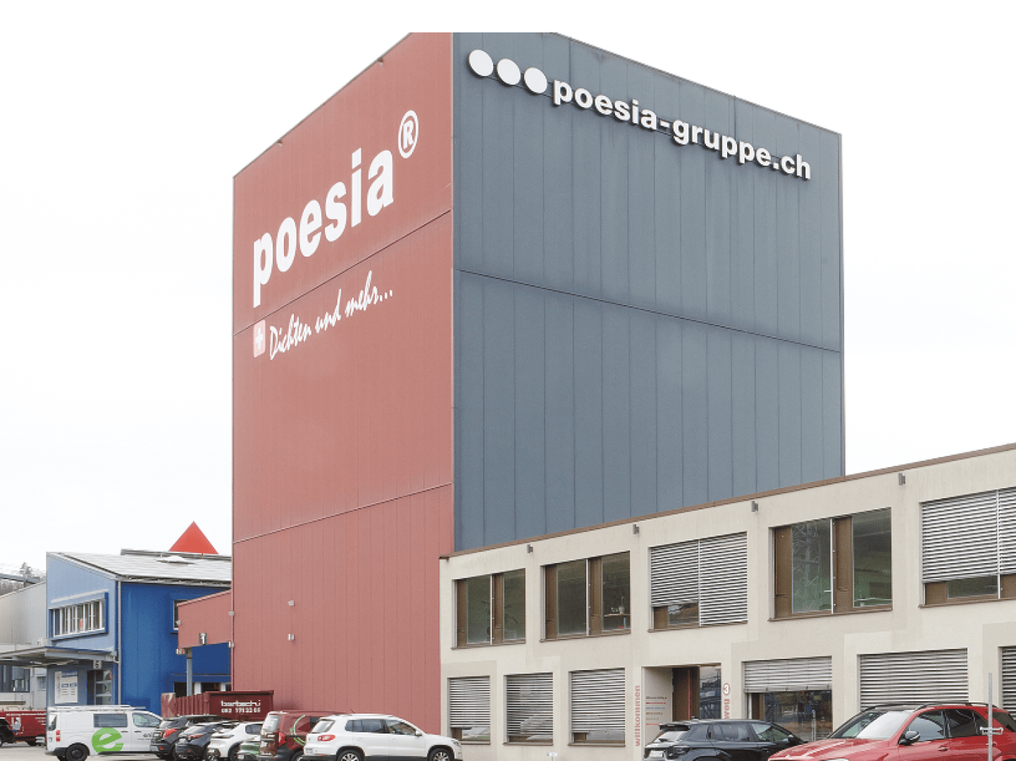 Ein modernes Industriegebäude mit einem hohen rot-grauen Turm mit der Aufschrift "poesia" und "poesia-gruppe.ch", umgeben von parkenden Autos und angrenzenden Gebäuden. Der Himmel ist bedeckt.
