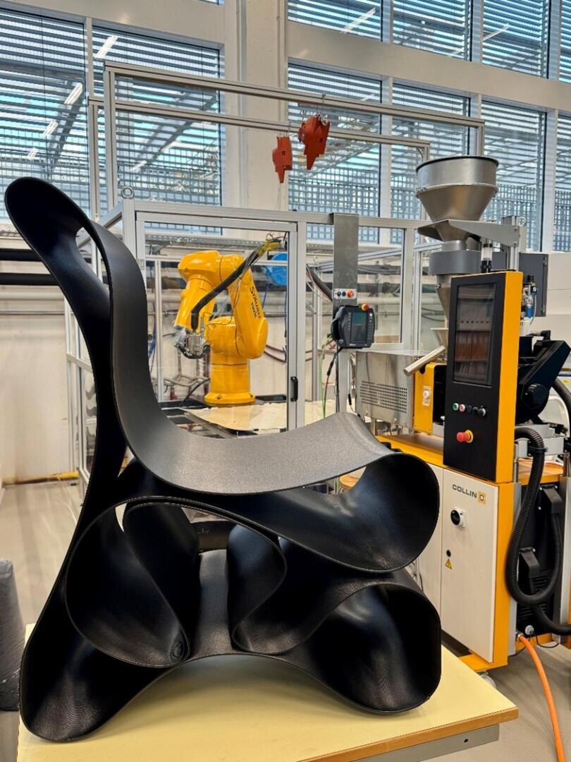 Ein skulpturaler schwarzer Stuhl mit geschwungenen, fließenden Formen sitzt vor einem gelben Industrieroboter und Maschinen in einer modernen, gut beleuchteten Fabrik- oder Laborumgebung.