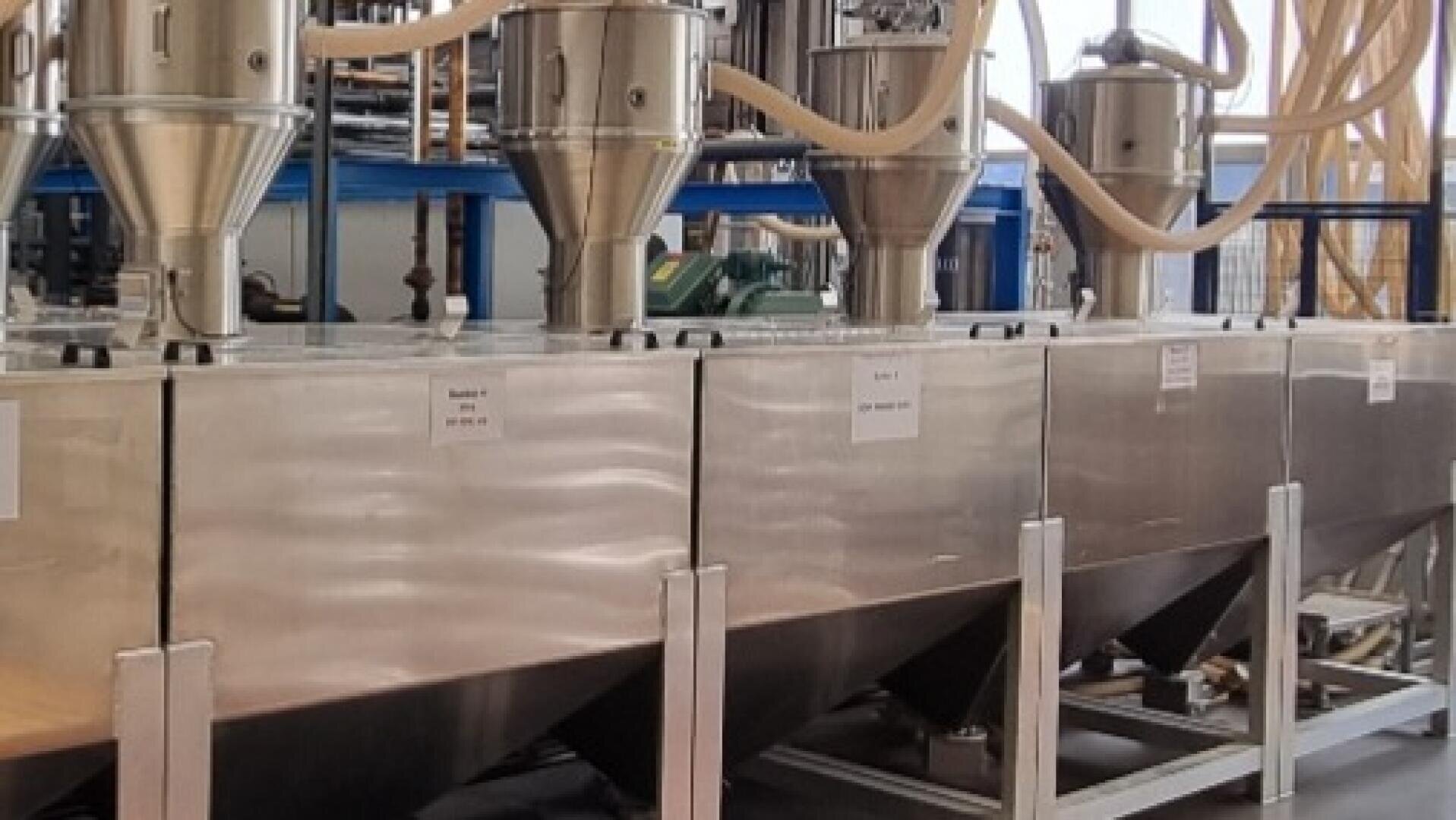 Eine Industriemaschine aus rostfreiem Stahl mit mehreren Trichtern und Rohren ist in einer Fabrikumgebung aufgestellt und wird wahrscheinlich für die Verarbeitung oder den Transport von Schüttgut verwendet. An der Anlage sind Etiketten und Rohre zu sehen.