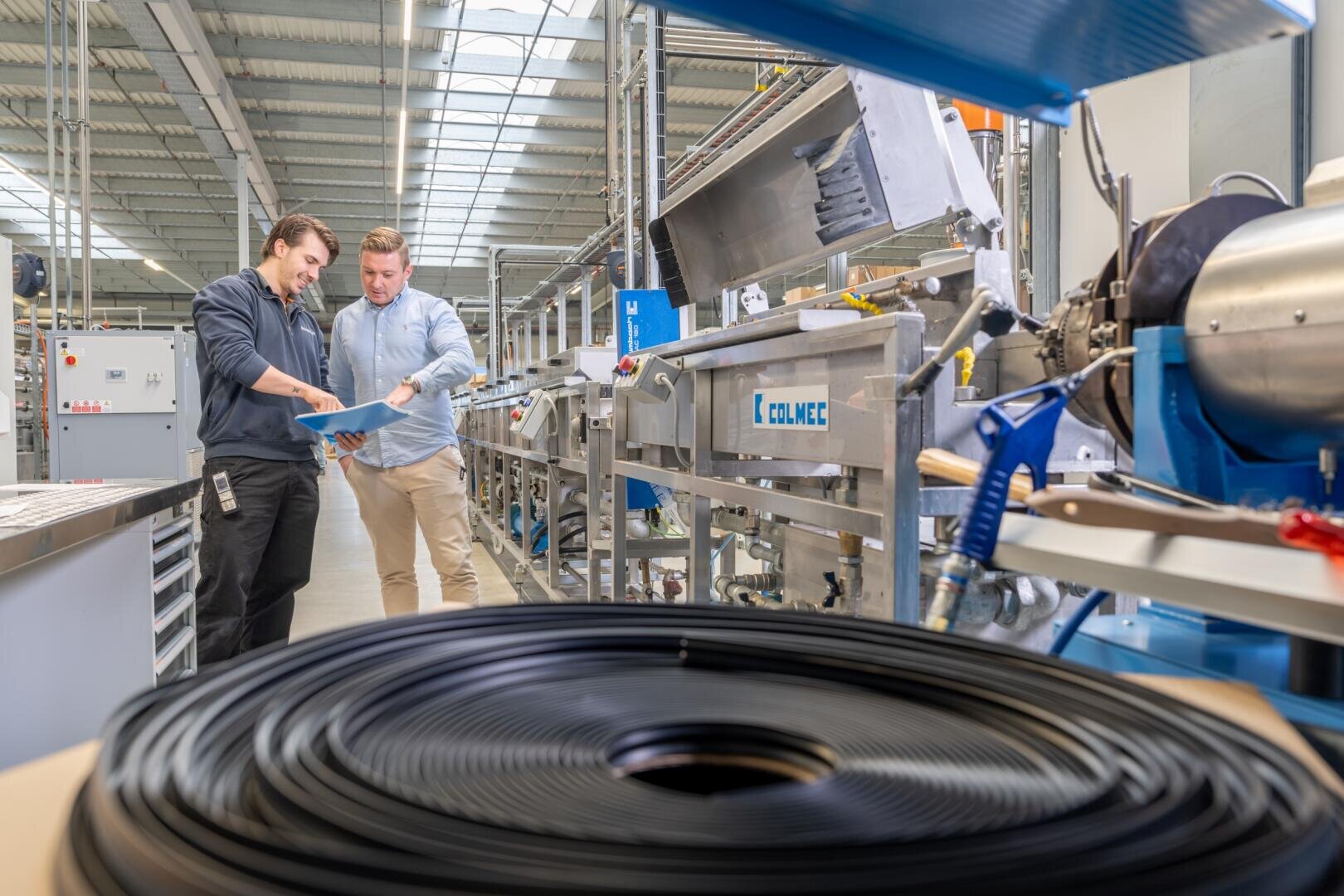 Zwei Männer stehen in einer Fabrik in der Nähe von Industriemaschinen und prüfen Dokumente. Im Vordergrund sind aufgerollte schwarze Kabel zu sehen, und der Arbeitsbereich ist gut beleuchtet, mit Metallgeräten und Regalen im Hintergrund.