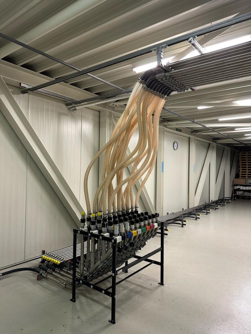 In einem Industrieraum mit hellen Wänden und einer Uhr an der gegenüberliegenden Wand wölbt sich eine Reihe flexibler Schläuche von der Decke herab und ist mit einem Metallgestell mit beschrifteten Ventilen verbunden.