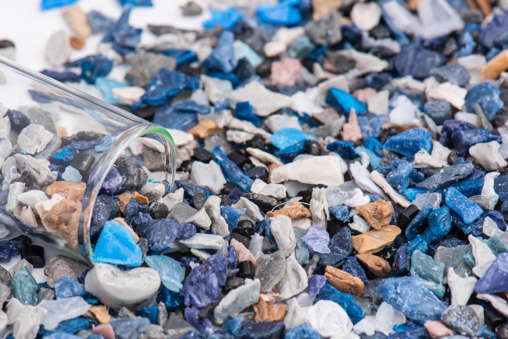Eine Nahaufnahme verschiedener kleiner, bunter Plastikfragmente, meist in Blautönen, die auf einer Fläche verstreut sind, auf der ein Klarglasbehälter steht, der zwischen den Stücken umkippt.