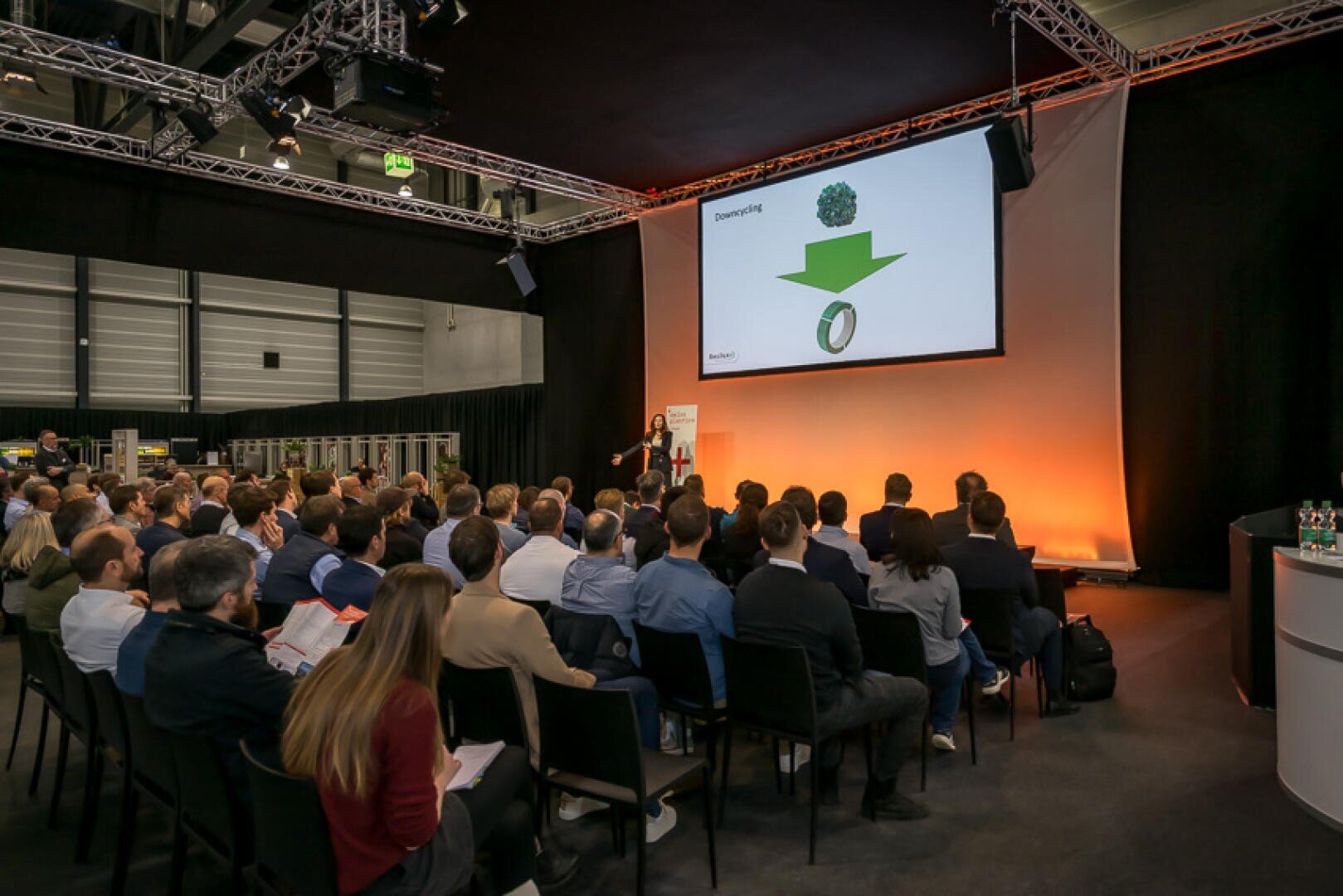 Ein Redner hält einen Vortrag vor einem sitzenden Publikum in einem modernen Konferenzraum. Auf einem großen Bildschirm wird eine Folie mit einem grünen Virussymbol, einem grünen Pfeil und einem medizinischen Armband angezeigt. Der Raum ist mit Oberlichtern und orangefarbenen Akzenten beleuchtet.