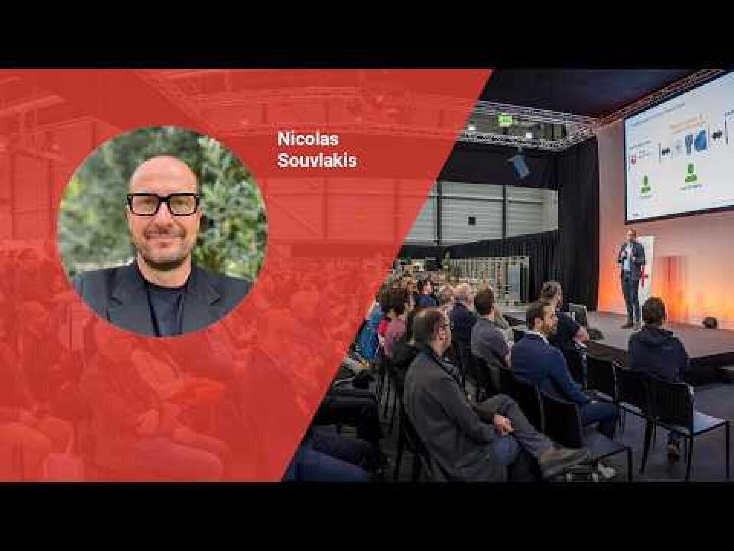 Ein Redner präsentiert auf der Bühne vor einem Publikum in einem modernen Veranstaltungsraum. Auf der linken Seite zeigt ein rotes Overlay das runde Foto eines Mannes mit Brille und den Namen Nicolas Souvlakis.