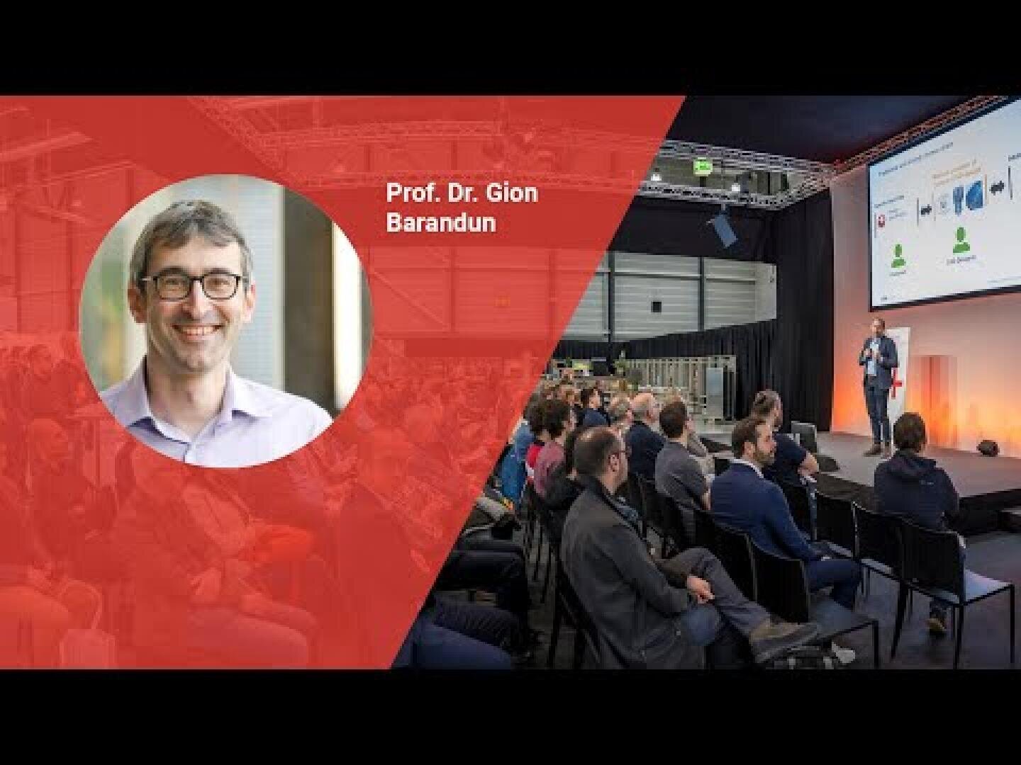 Ein großes Publikum hört einem Redner auf der Bühne bei einer Konferenz zu. Ein Foto von Prof. Dr. Gion Barandun und sein Name sind auf einem roten Overlay auf der linken Seite des Bildes zu sehen.