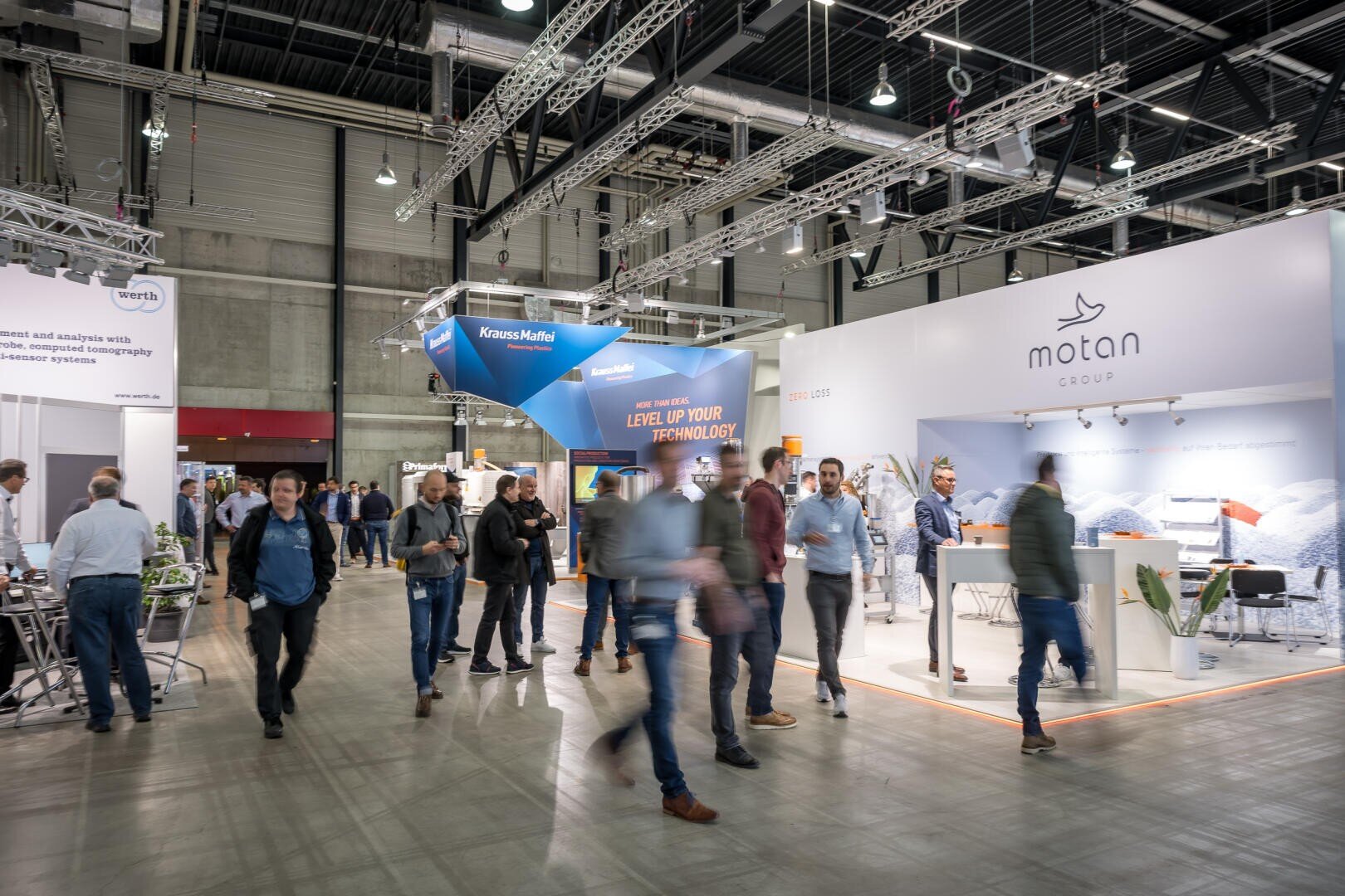Les gens traversent un hall d'exposition très animé, avec des stands de différentes entreprises, dont KraussMaffei et le groupe Motan, qui présentent leurs produits et leurs bannières sous des projecteurs très lumineux.