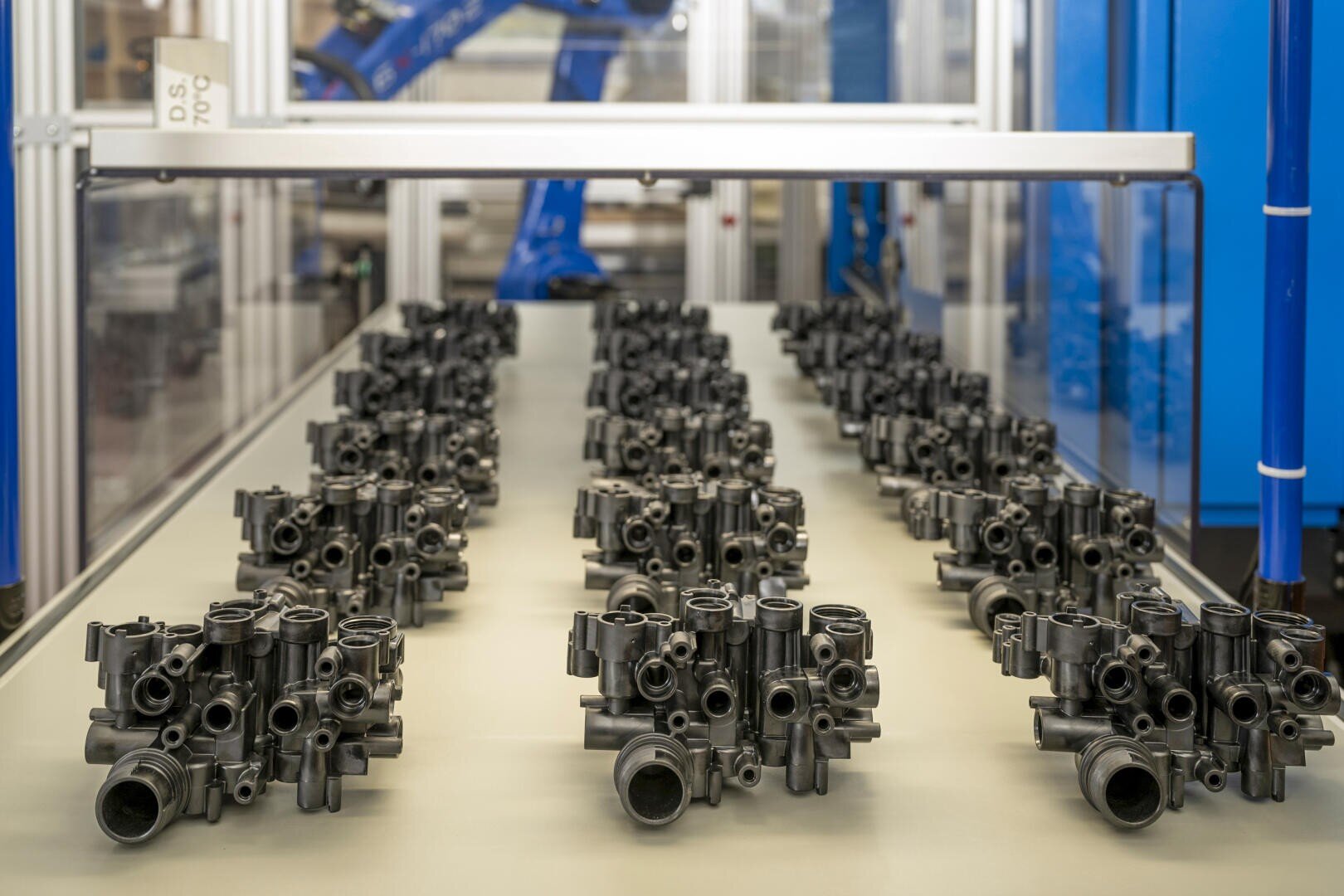 Reihen identischer schwarzer Maschinenteile aus Metall sind in einer industriellen Umgebung fein säuberlich auf einem Förderband angeordnet, im Hintergrund sind blaue Robotermaschinen zu sehen.