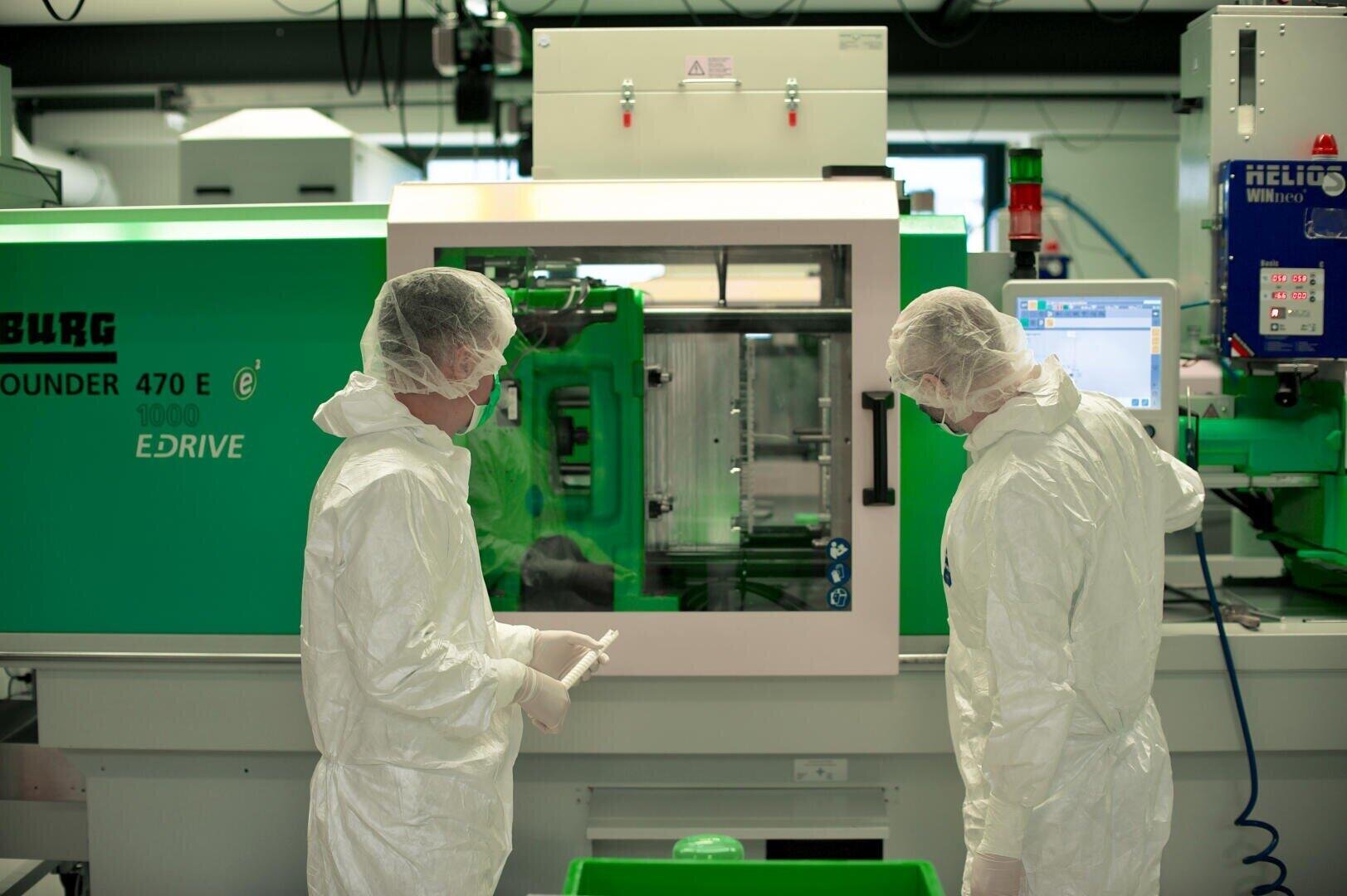 Deux personnes vêtues de combinaisons de protection blanches, de résilles et de masques travaillent dans un laboratoire ou une usine avec une machine industrielle verte, surveillent son fonctionnement et utilisent les appareils.