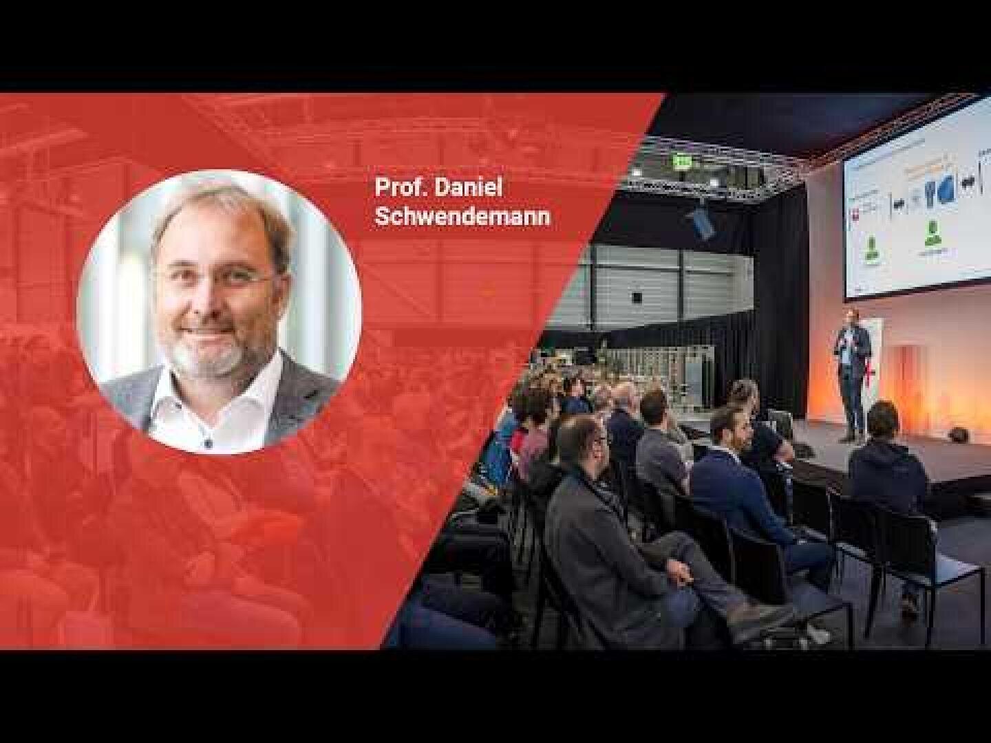 Ein Mann spricht auf der Bühne zu einem Publikum in einem modernen Konferenzraum. Auf der linken Seite zeigt eine rote Grafikeinblendung ein Porträt und den Namen von Professor Daniel Schwendemann. Im Hintergrund wird eine Präsentationsfolie projiziert.
