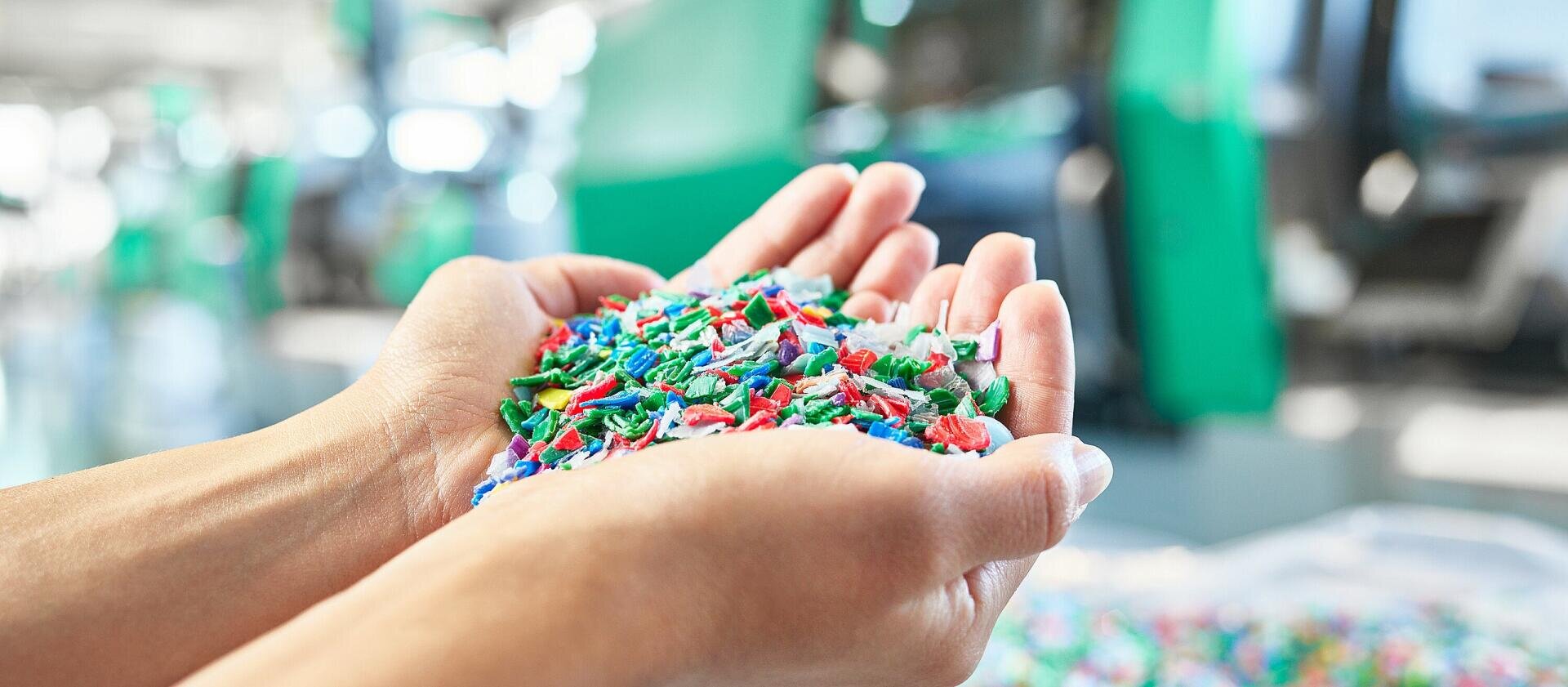 Ein Paar Hände hält einen Haufen bunter Plastikkügelchen oder -fragmente vor einem unscharfen industriellen Hintergrund.