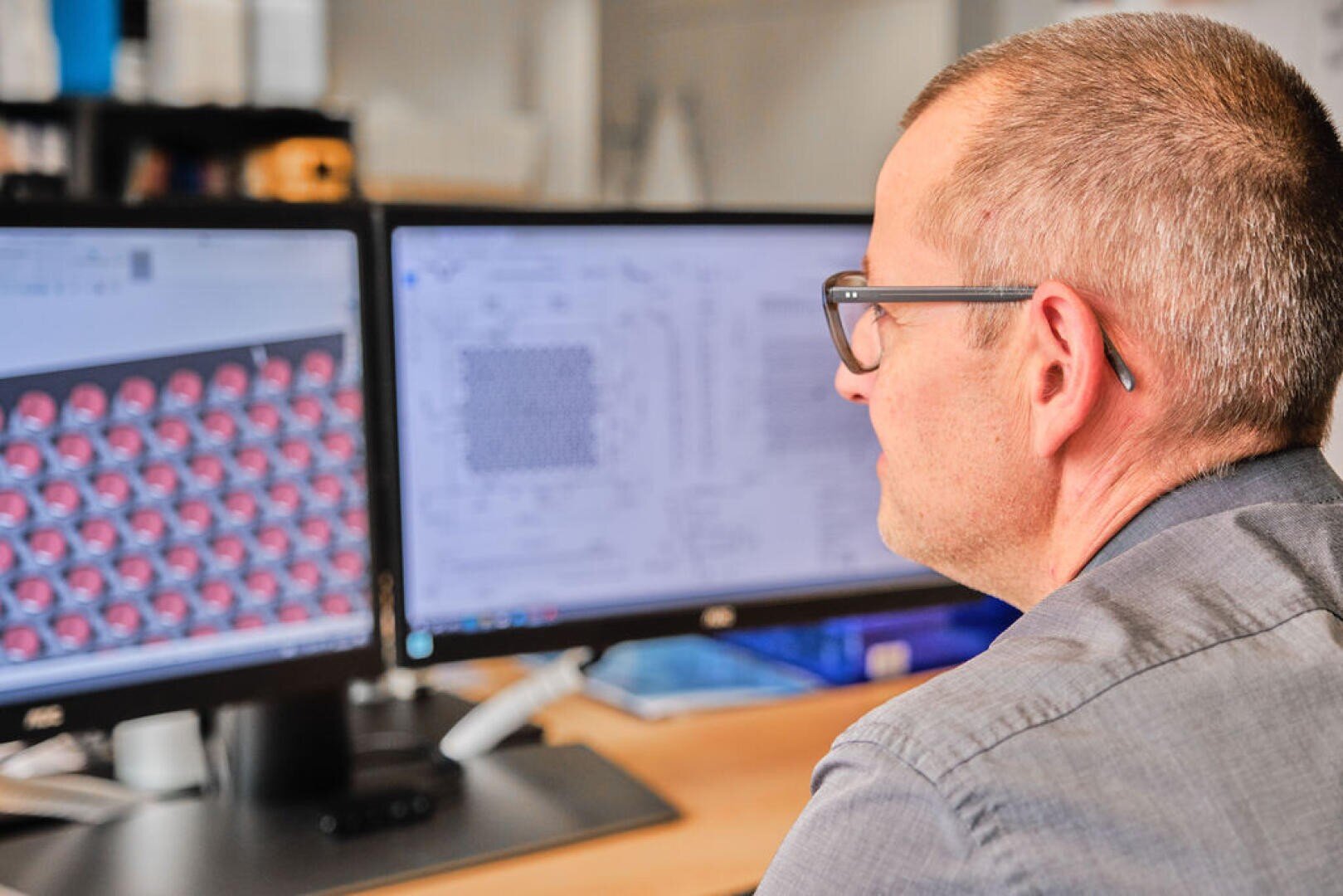 Eine Person, die eine Brille trägt, sitzt an einem Schreibtisch und arbeitet an zwei Computermonitoren, auf denen technische und 3D-Modellierungsentwürfe in einer Büroumgebung angezeigt werden.