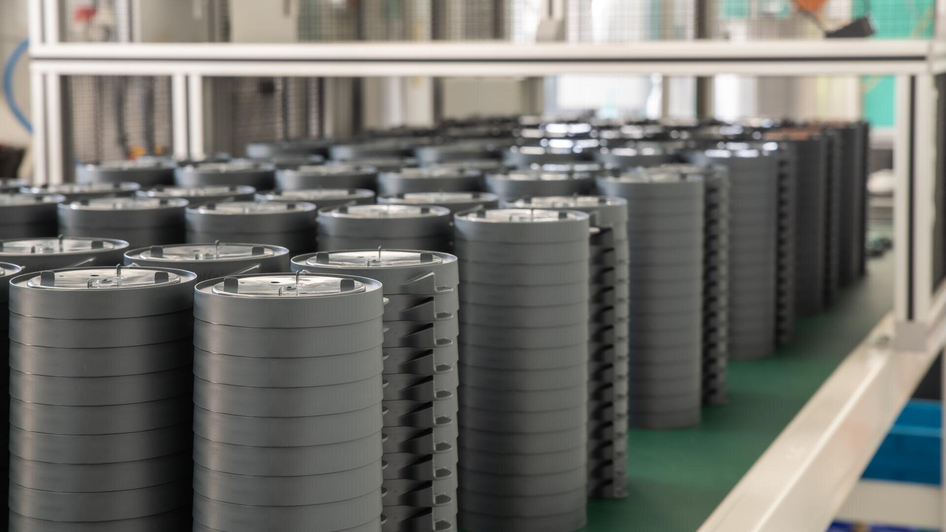 Stapel von zylindrischen Lithium-Ionen-Batteriezellen sind in Reihen auf einer grünen Fläche in einer modernen Produktionsanlage angeordnet. Im Hintergrund sind Industrieanlagen und eine saubere, organisierte Umgebung zu sehen.