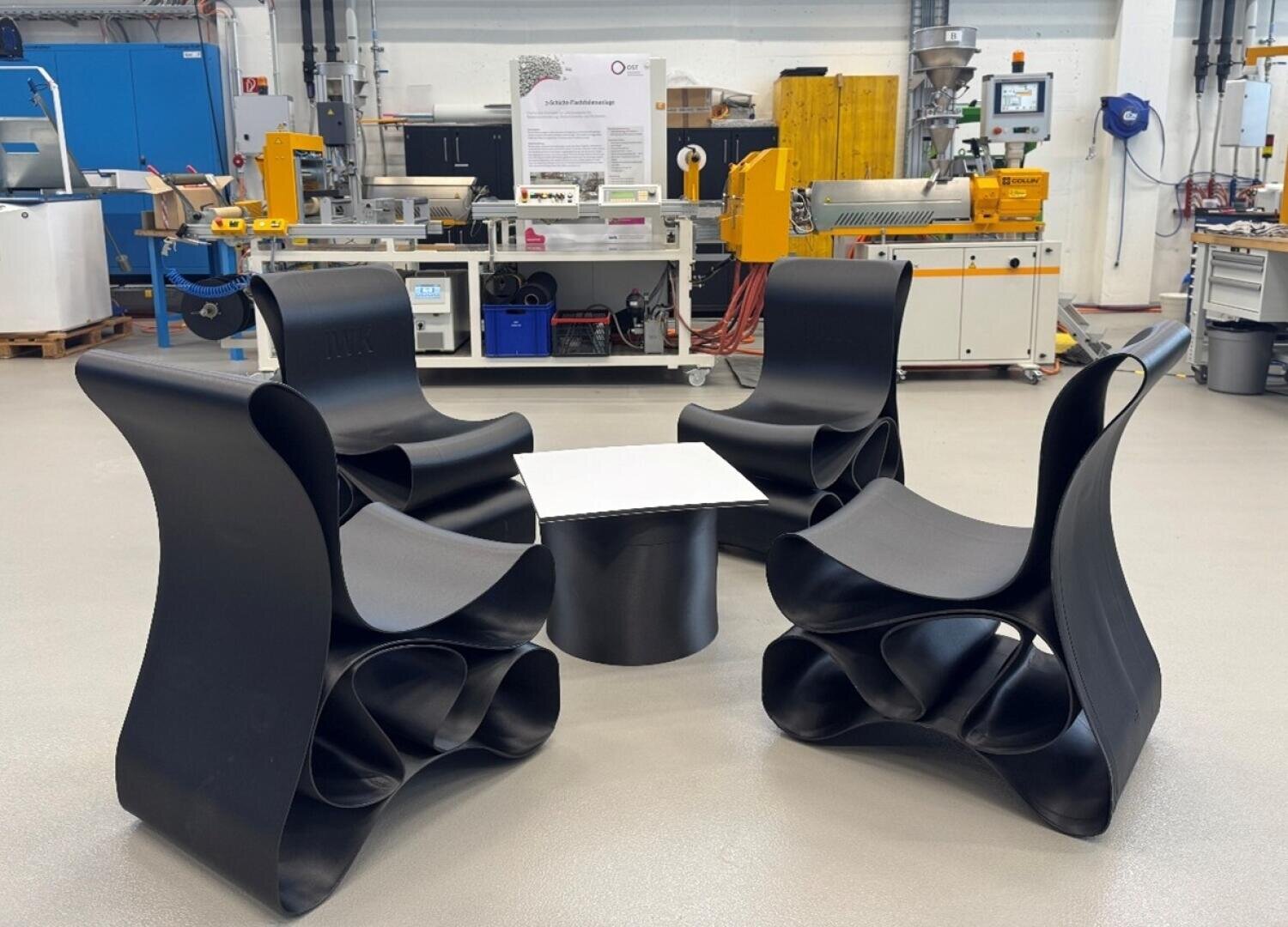 Vier moderne schwarze Stühle mit geschwungenen, skulpturalen Formen sind um einen kleinen quadratischen Tisch in einer hellen Werkstatt voller industrieller Geräte und Maschinen angeordnet.