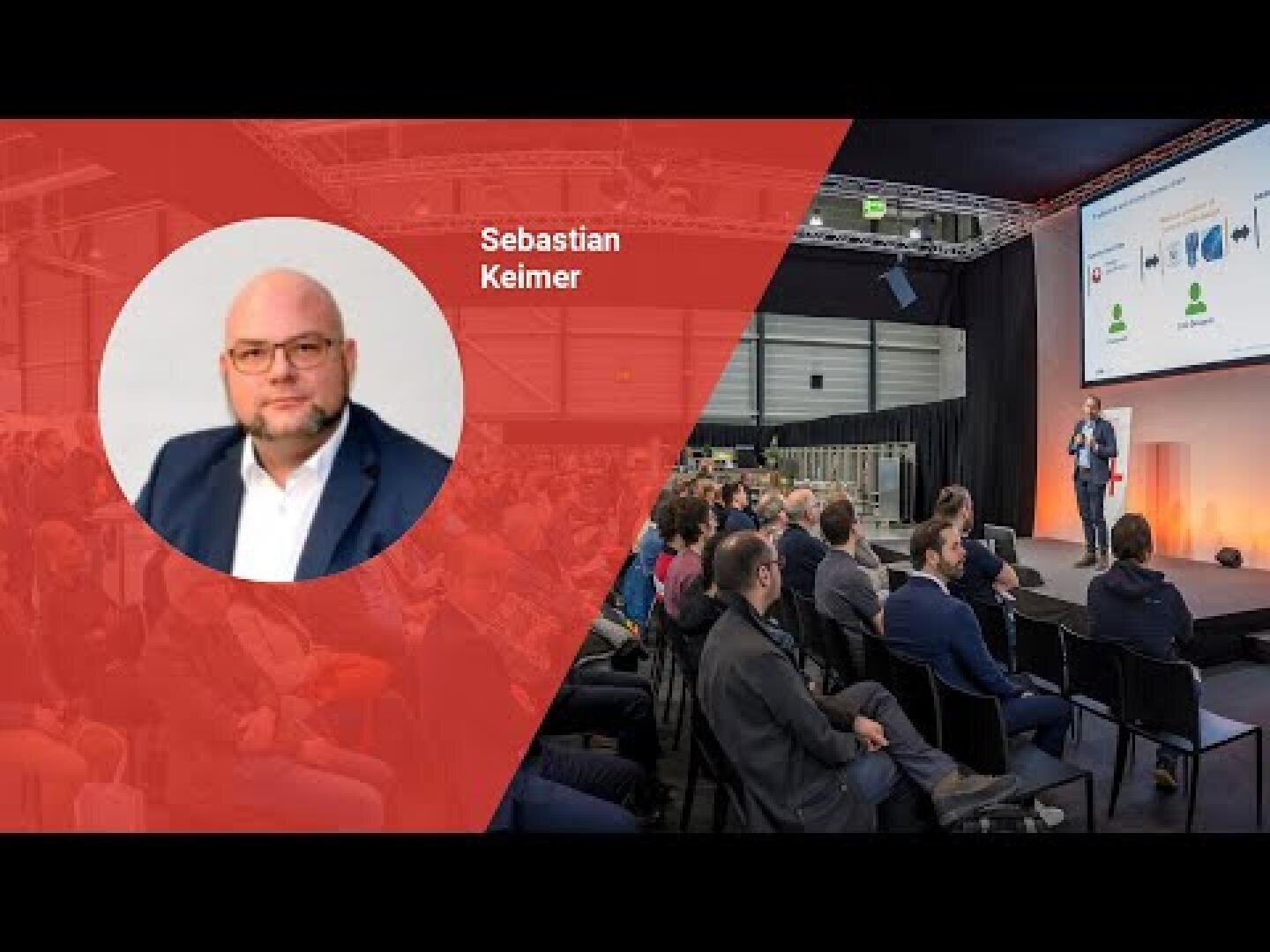 Un orateur donne une conférence sur scène devant un public assis en rangées. Sur le côté gauche, la photo d'un homme chauve, portant des lunettes et une barbe, portant la mention Sebastian Keimer, s'affiche en rouge.