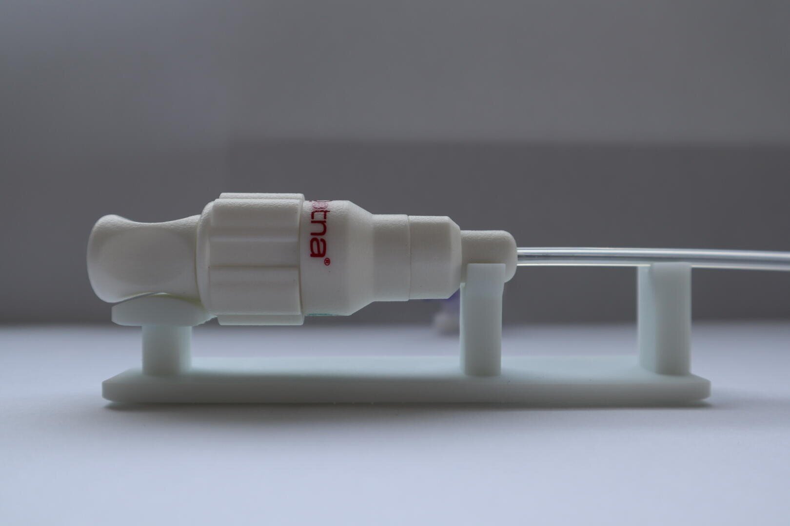 Gros plan sur un appareil médical muni d'un raccord en plastique blanc fixé à un tuyau transparent et reposant sur un support blanc. Le raccord est partiellement étiqueté en rouge et photographié sur un fond gris uni.