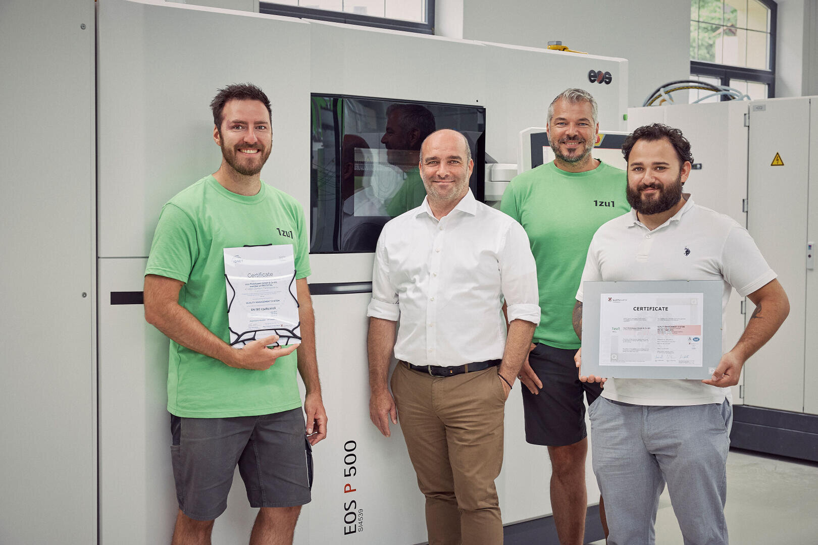 Quatre hommes se tiennent devant une machine EOS P 500. Deux hommes en chemise verte tiennent des certificats, tandis que les deux autres en chemise blanche se tiennent à côté. Tous sourient dans un environnement industriel ou de laboratoire.