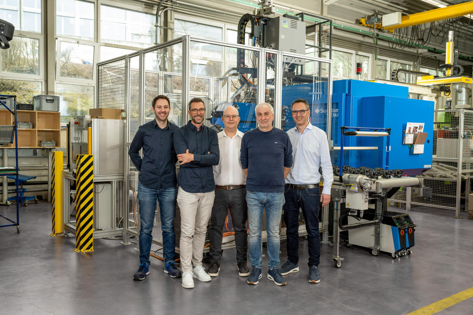 Fünf Männer stehen lächelnd nebeneinander in einer hellen Industriehalle mit Maschinen und Geräten im Hintergrund. Die Umgebung zeichnet sich durch große Fenster und organisierte Arbeitsbereiche aus.