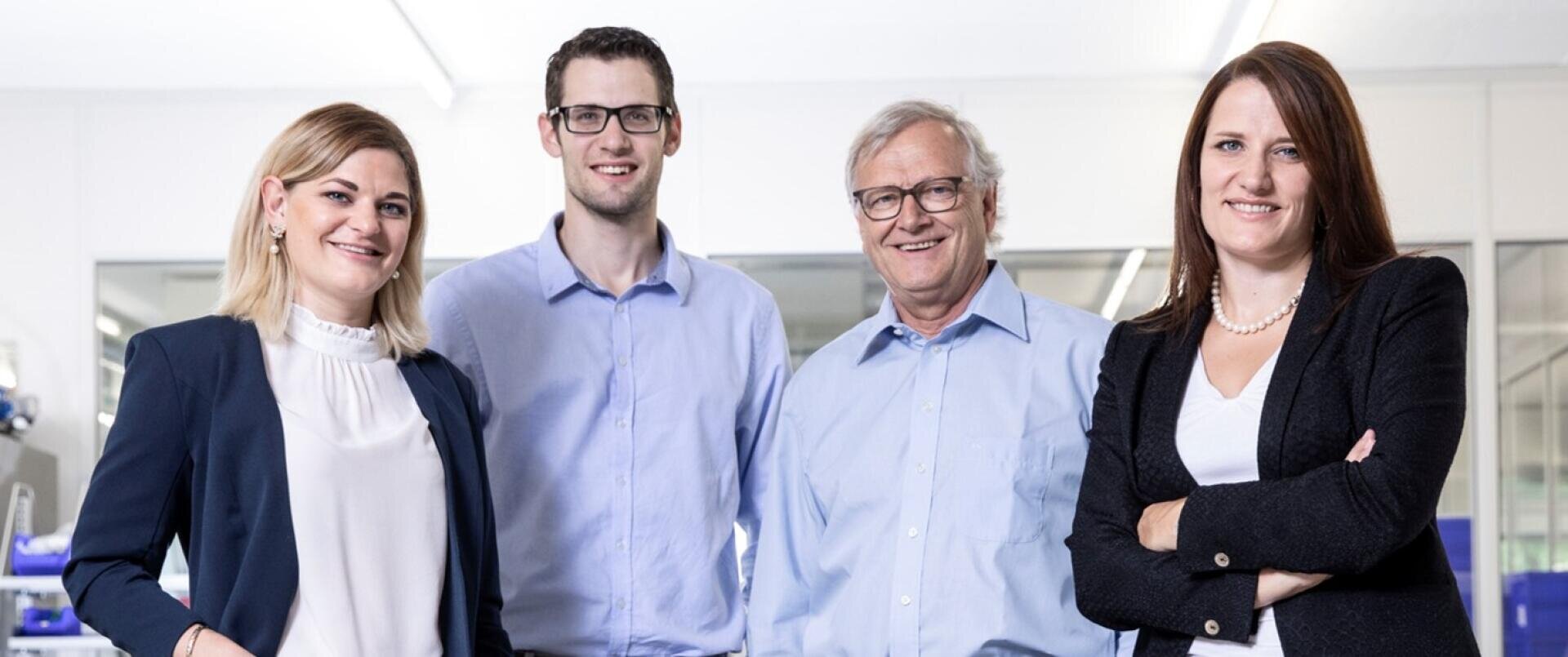 Quatre adultes - deux femmes et deux hommes - se tiennent ensemble dans une maison et sourient à la caméra. Ils sont habillés en vêtements d'affaires, ce qui laisse supposer un environnement professionnel ou un bureau.