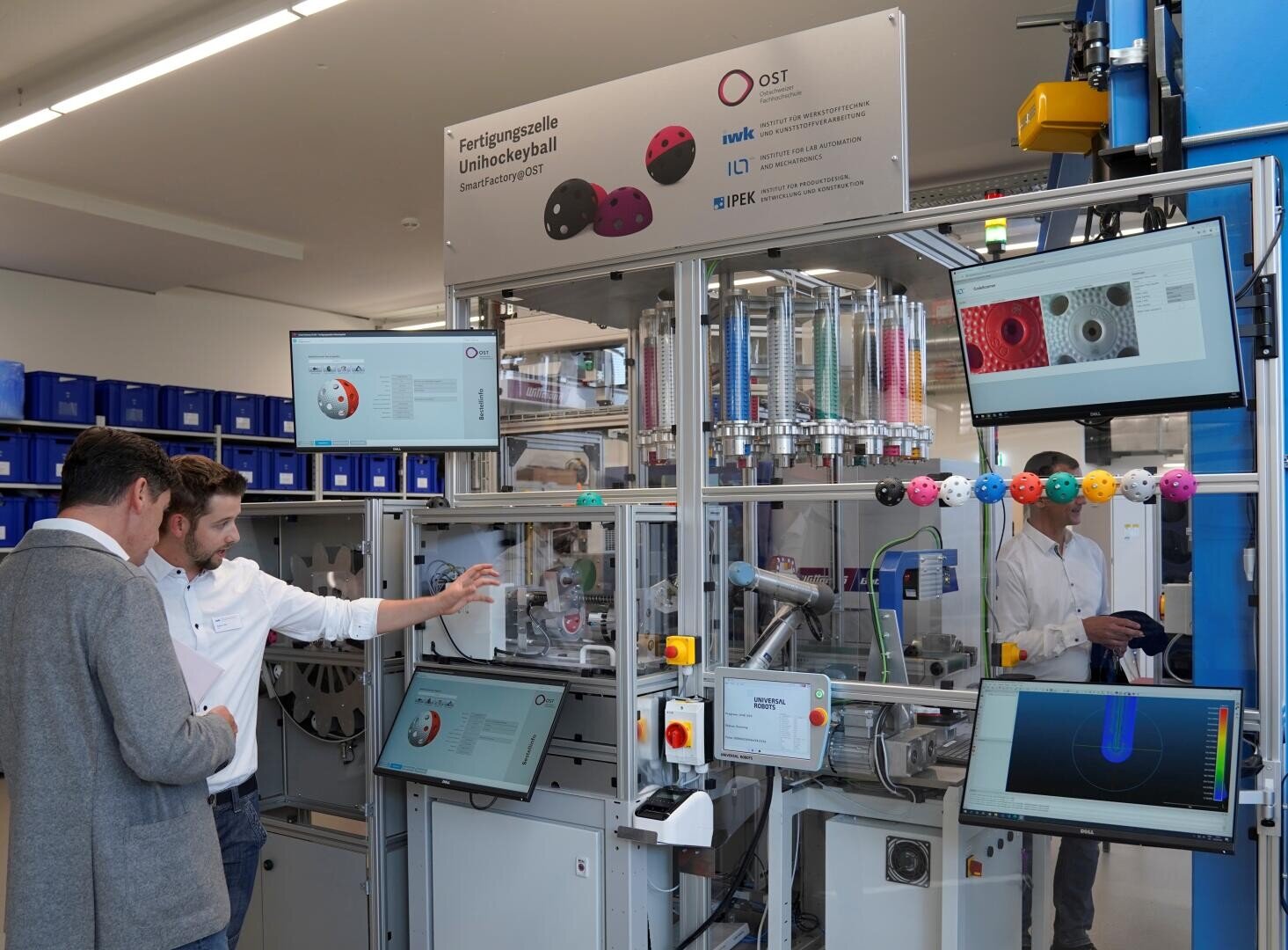 Zwei Männer stehen vor einer komplexen Industriemaschine mit mehreren Bildschirmen, Diagrammen und bunten Plastikkugeln; eine weitere Person ist im Hintergrund durch Glas sichtbar. Technische Geräte und Lagerkästen säumen den Raum.