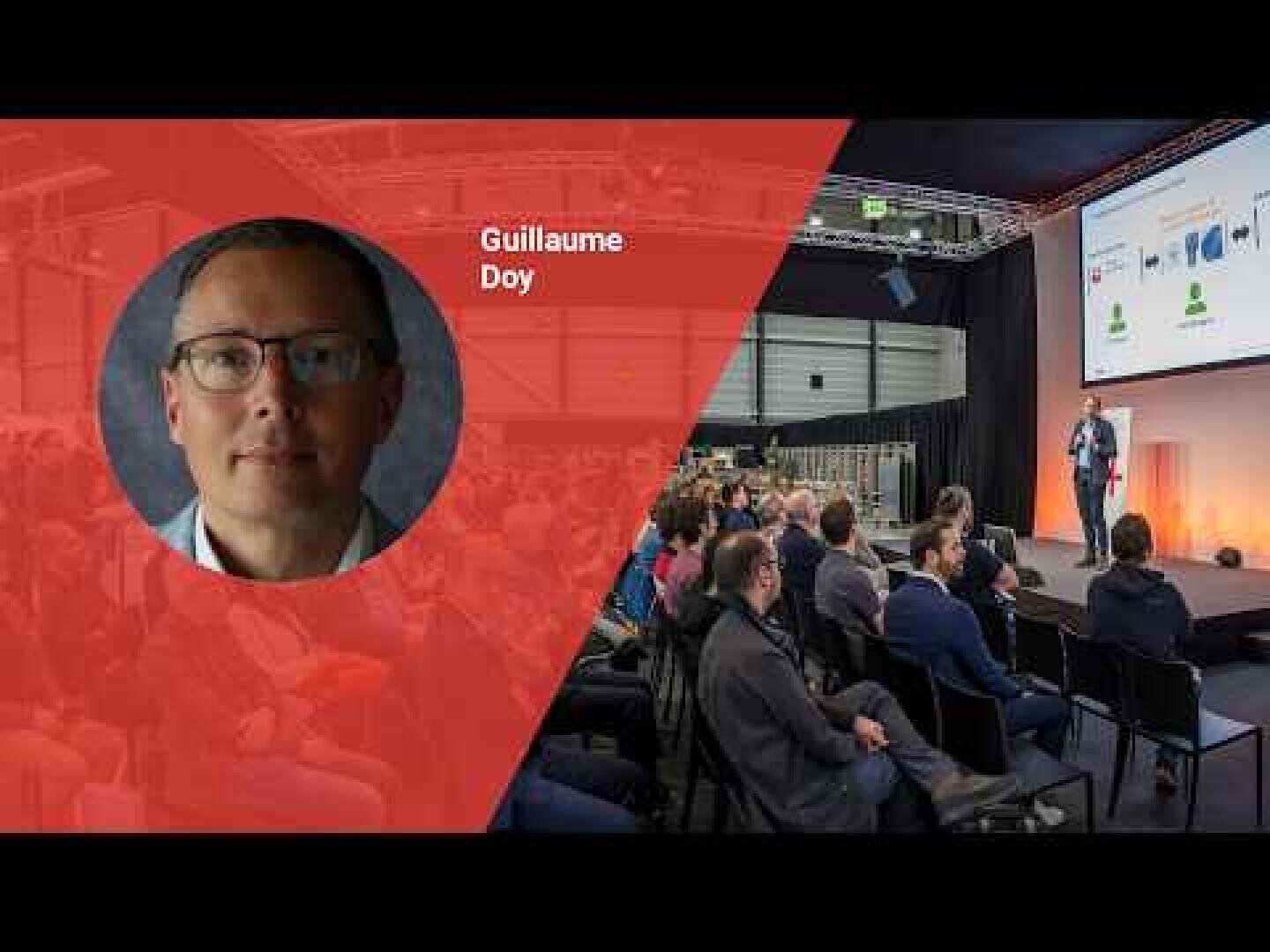 Ein Mann steht auf der Bühne und spricht zu einem Publikum, das in einem Konferenzraum sitzt. Auf der linken Seite zeigt eine überlagerte rote Grafik den Kopf eines Mannes namens Guillaume Doy.